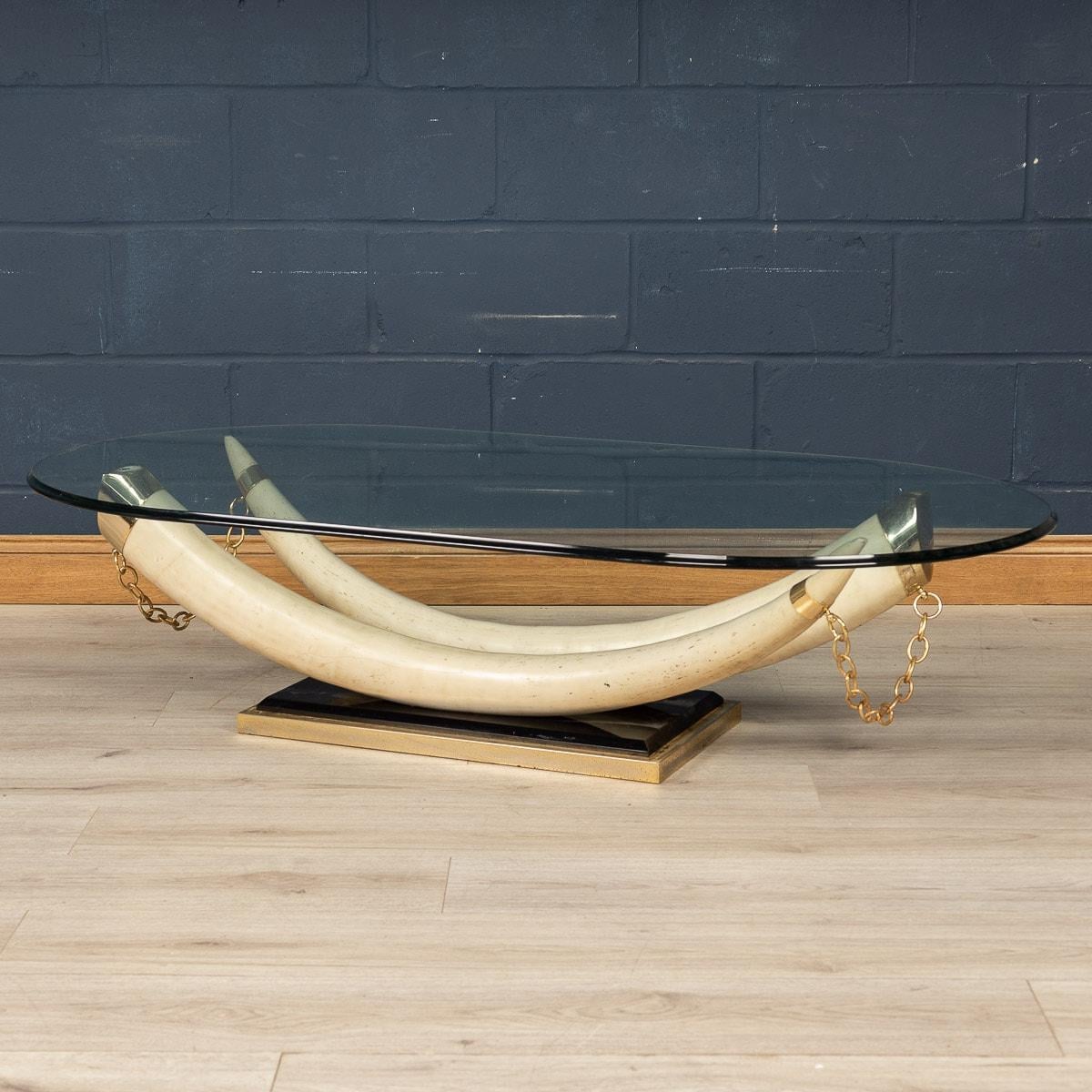 A striking French made coffee table in the early 1970s. The faux elephant tusks are made entirely out of resin and connected by a gold coloured chain over a black rectangular base with a brass surround. The tusks support an oval bevelled glass, all