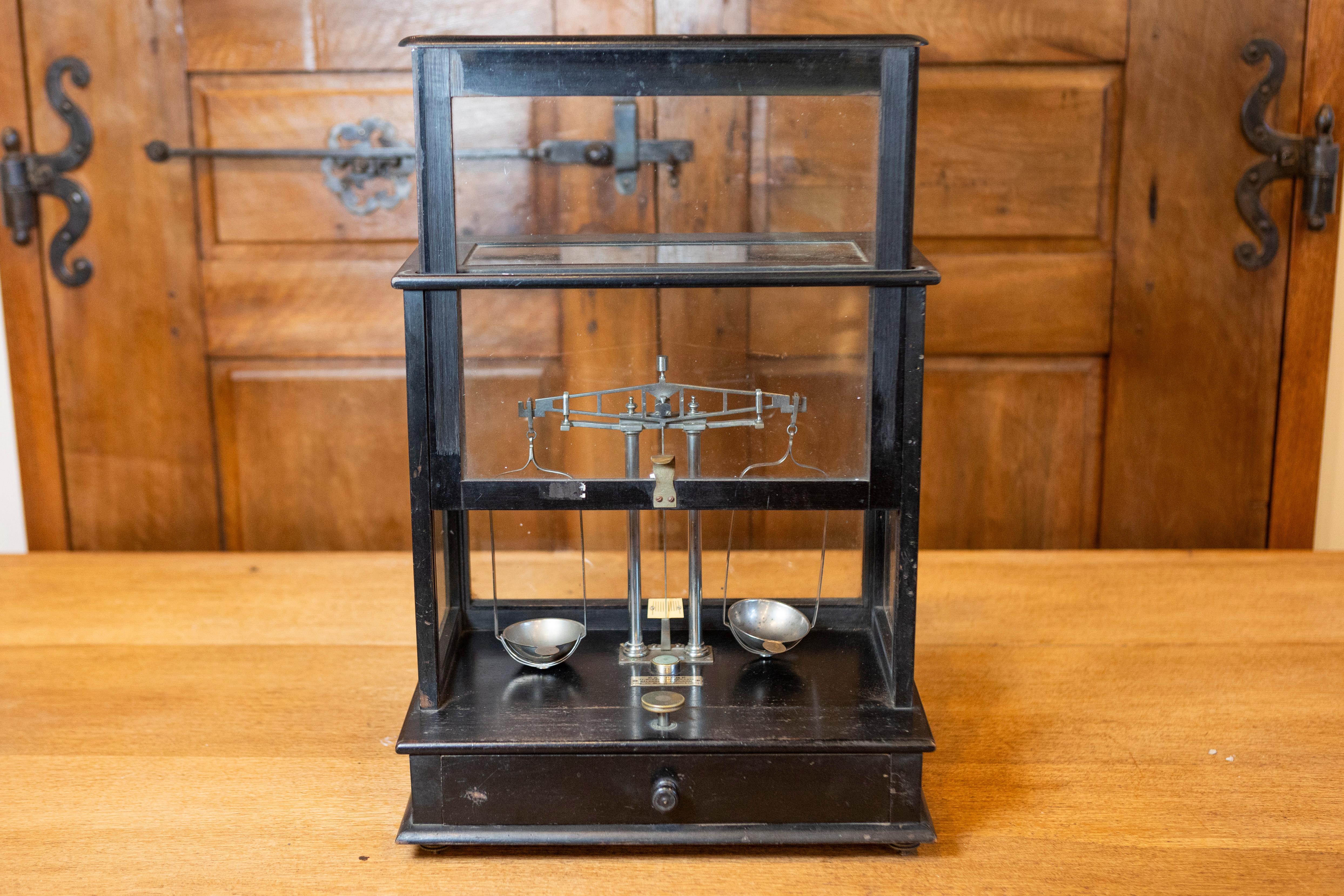 Cette balance française du XXe siècle encastrée dans du verre est une pièce fonctionnelle et décorative qui reflète un mélange d'artisanat et de design pratique. La balance est logée dans un cadre en bois noir avec des panneaux en verre sur tous les