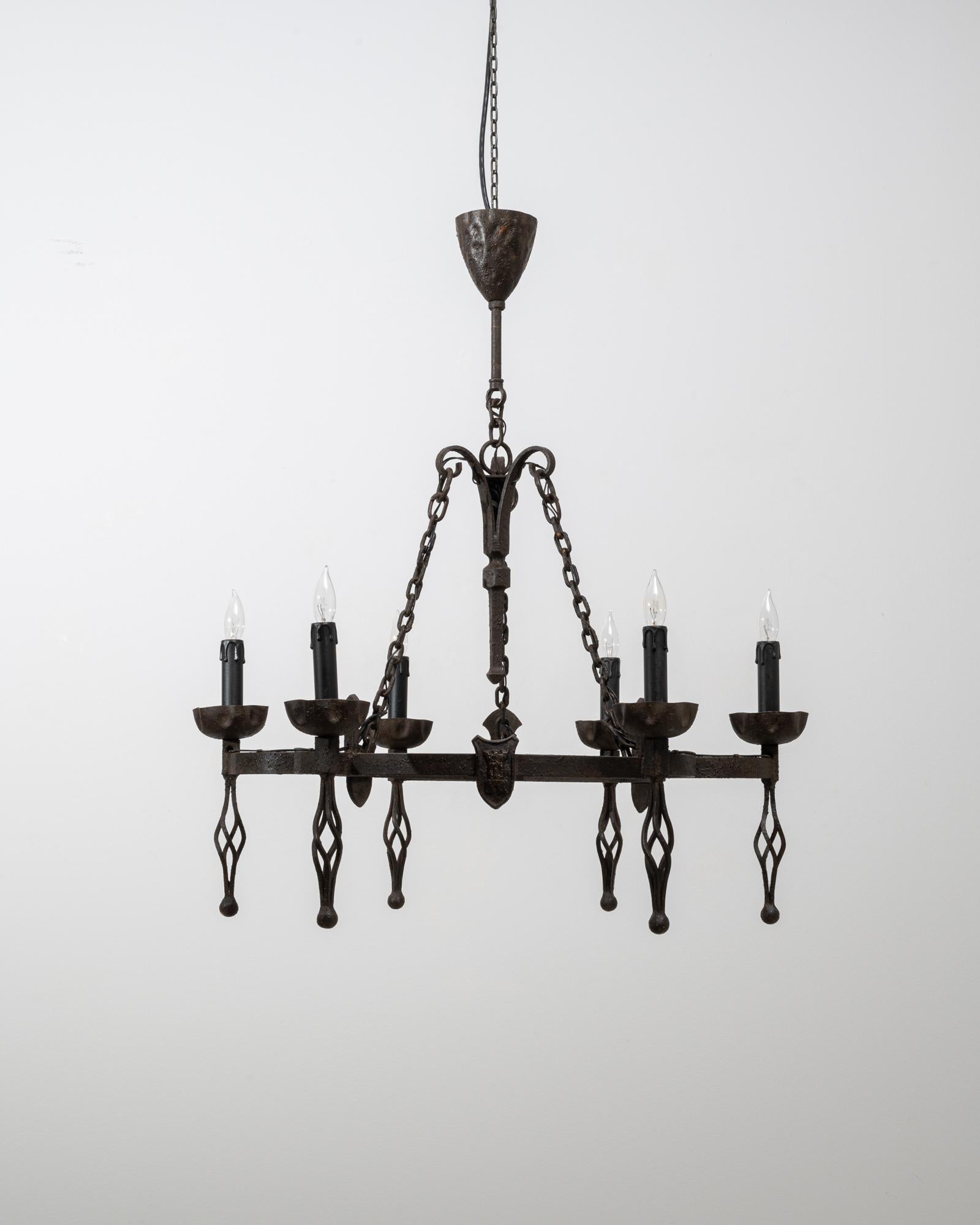 Esta araña de hierro forjado del renacimiento gótico francés del siglo XX muestra la presencia dramática y arquitectónica característica del estilo. Fabricada con un robusto armazón rectangular de hierro, tiene seis luces tipo vela sostenidas por