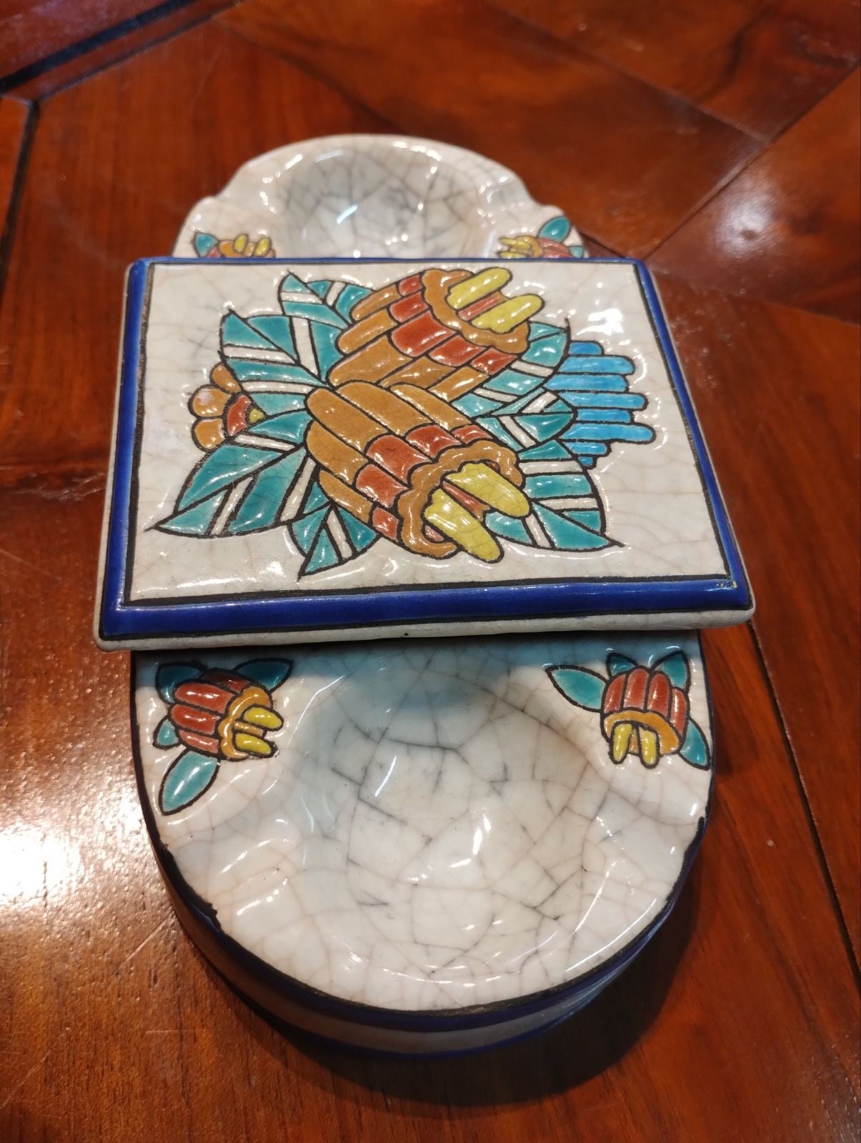 Hand painted oval ceramic ashtray with small covered part in the middle. The top of it is decorated with flowers and leaves in bright colours. There are numerous stamps and numbers of the edition at the bottom. Very good condition with no