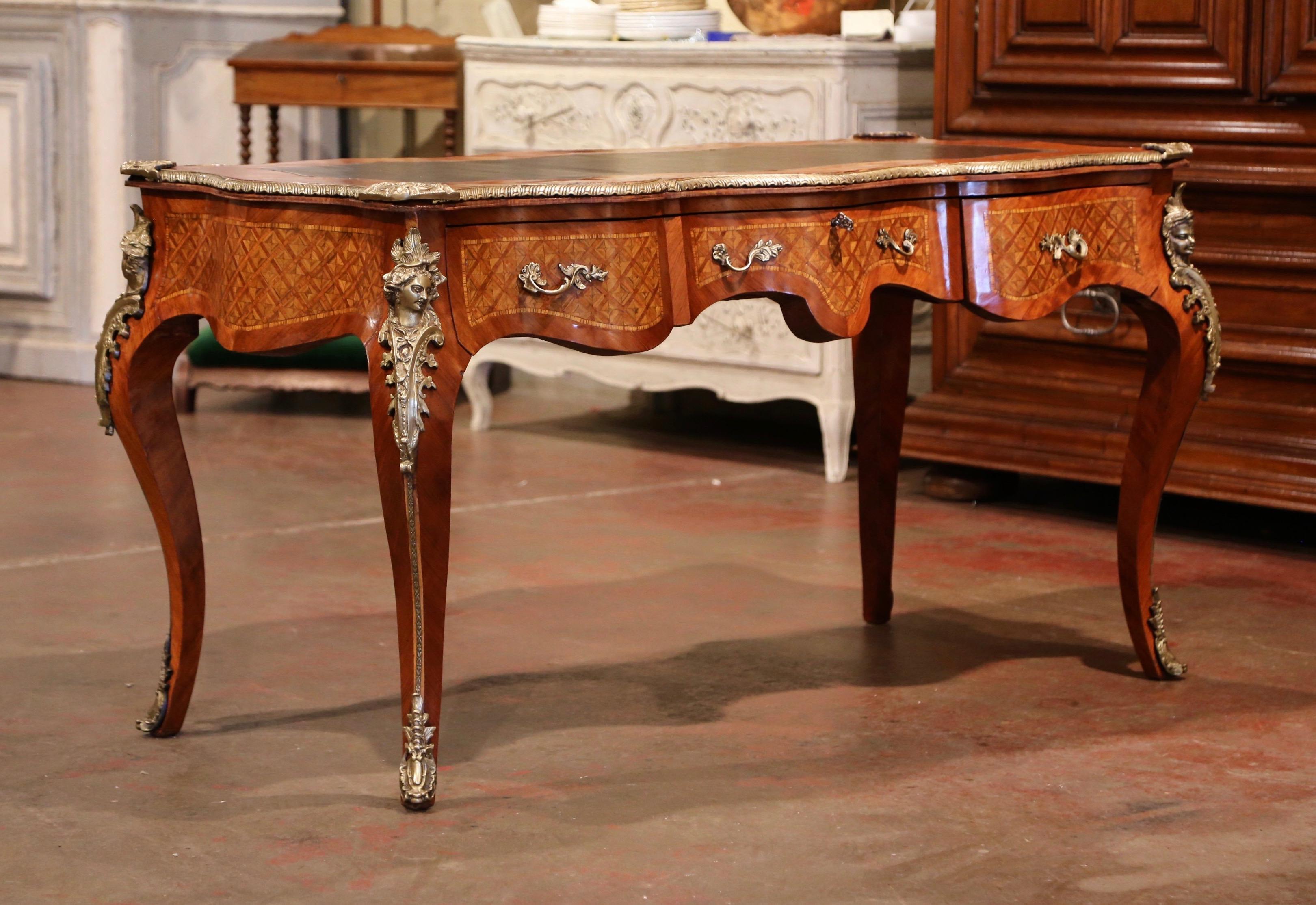 Created in France circa 1920, this elegant antique desk stands on cabriole legs decorated with bronze figural ormulu mounts at the shoulders and ending with sabots over the feet. The writing table features three serpentine drawers across the front