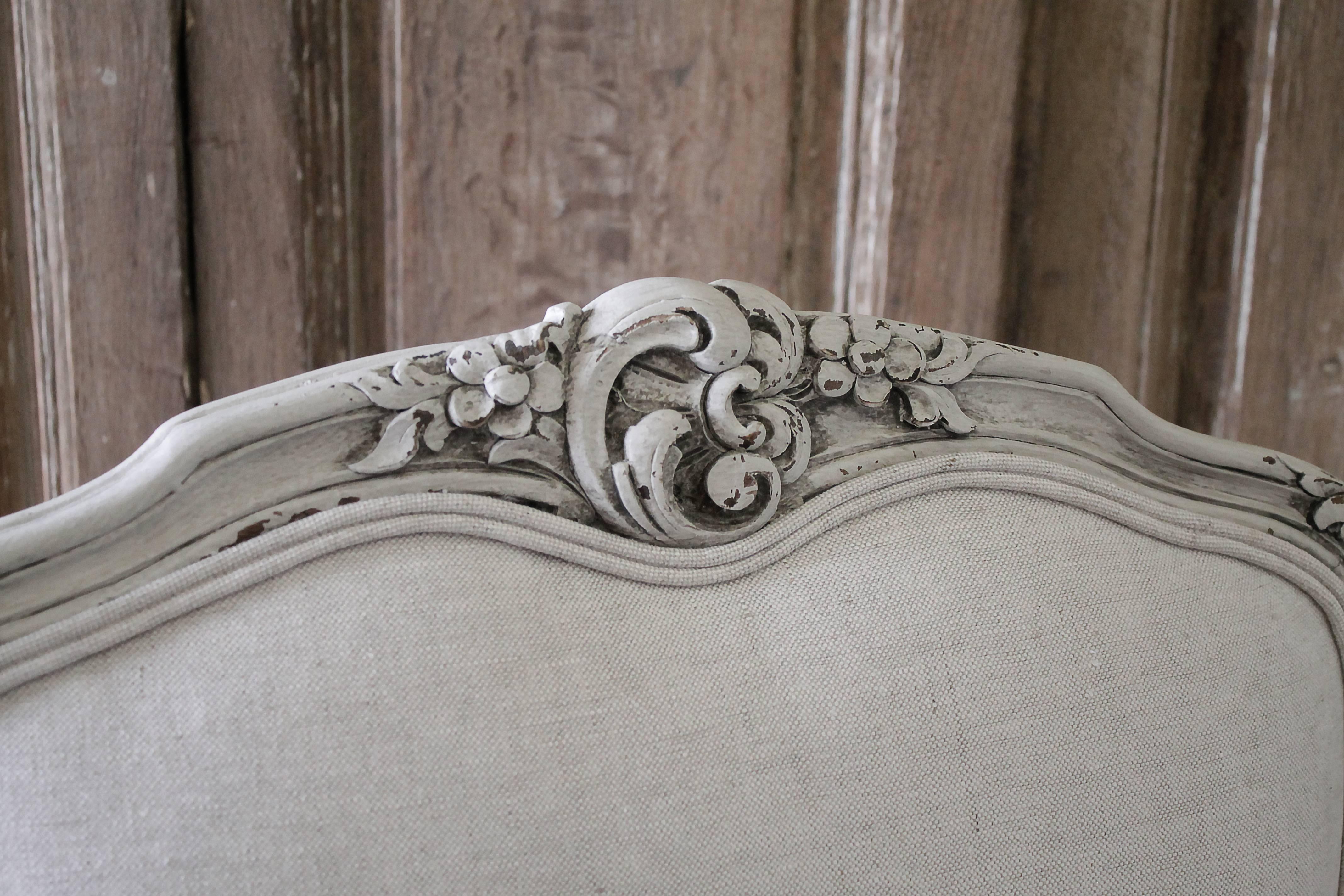 20th century French armchair upholstered in Belgian linen Painted in our oyster white, which is a true off-white with subtle distressed edges, and finished with an antique glazed patina. A cartouche carving with trailing floral vines are carved at
