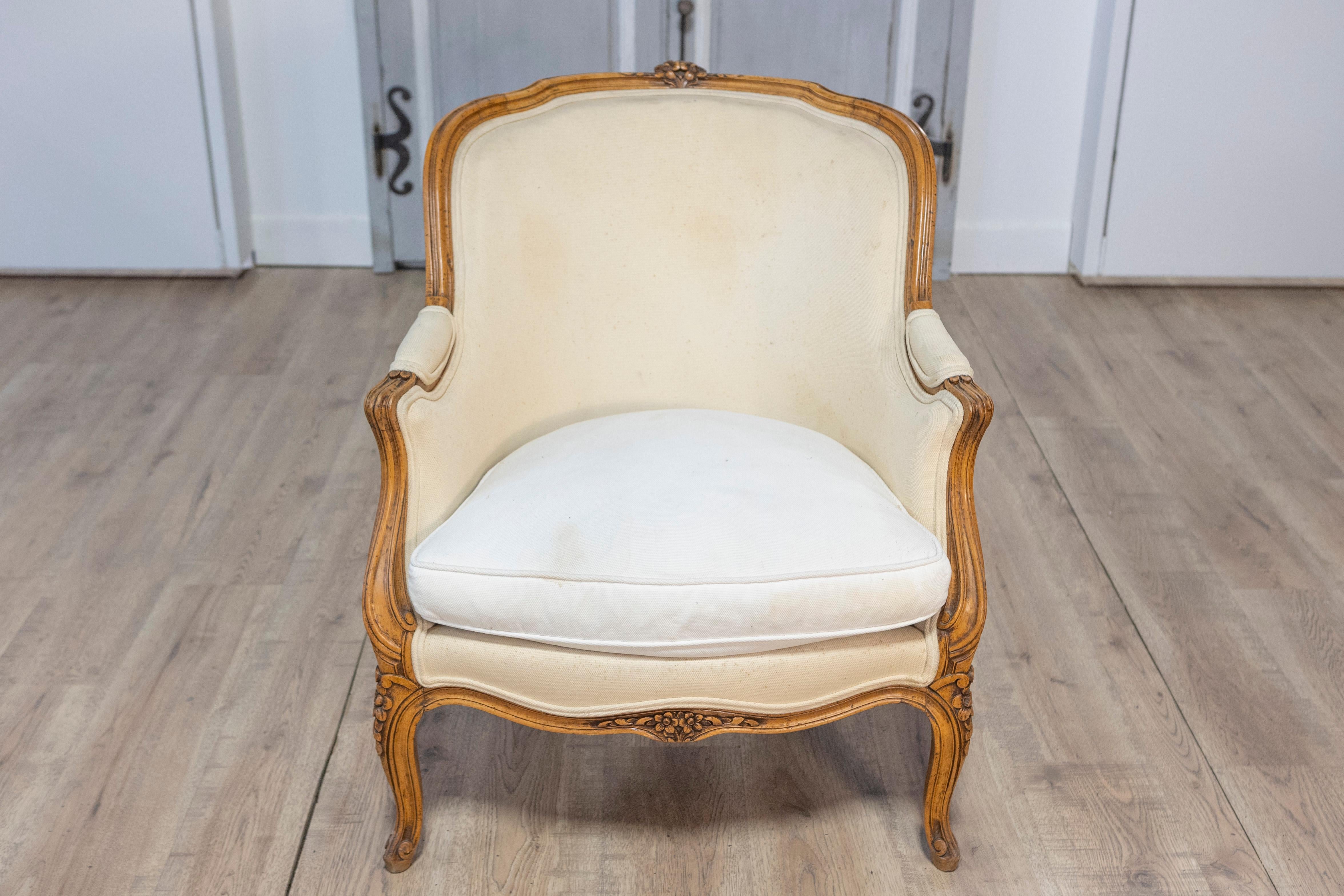 This 20th-century French bergère is an elegant representation of the Louis XV style, known for its graceful curves and refined ornamentation. The frame of the chair is crafted from carved wood, likely beech or walnut, and is finished in a warm,