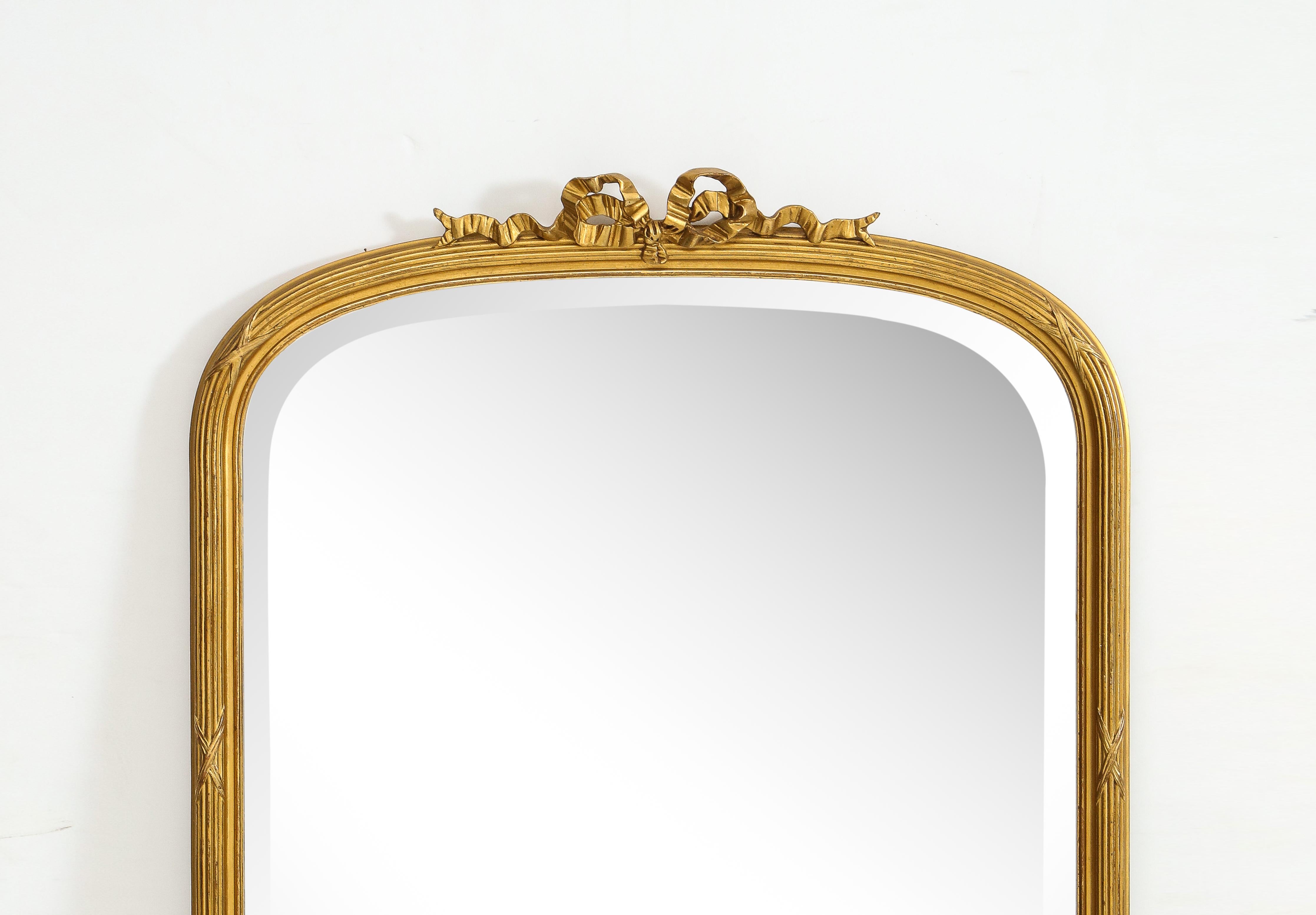20th Century French Louis XVI style gilt and gesso over-mantel mirror. The reeded rounded frame features a pierced bow and ribbon crest at the top, with a wide beveled glass plate.