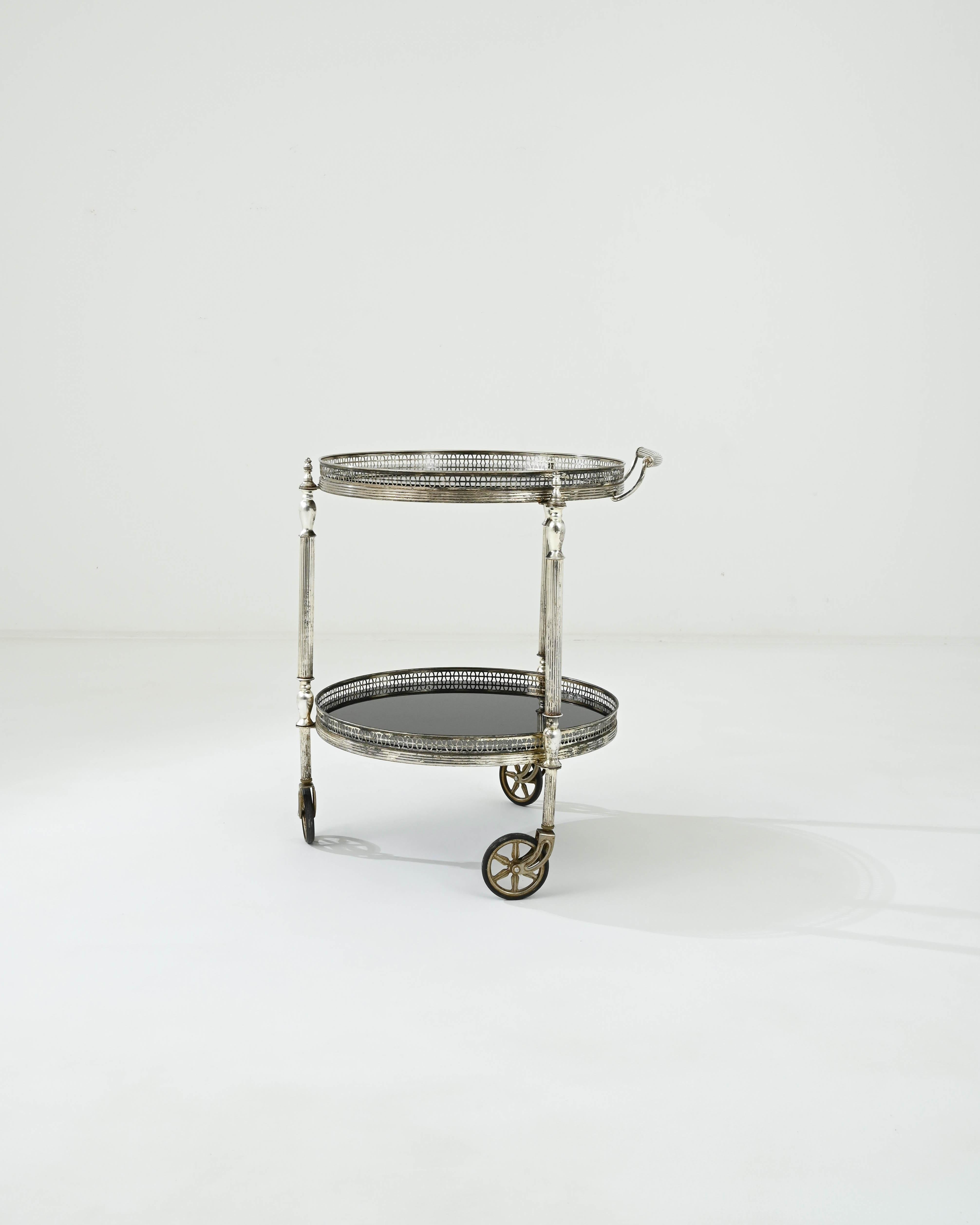 A metal and glass bar cart created in 20th Century France. This dazzling art deco inspired bar cart glitters with an opulent brilliance. The metal that composes its structure has been slowly covered in a compelling patina, subtly indicating its