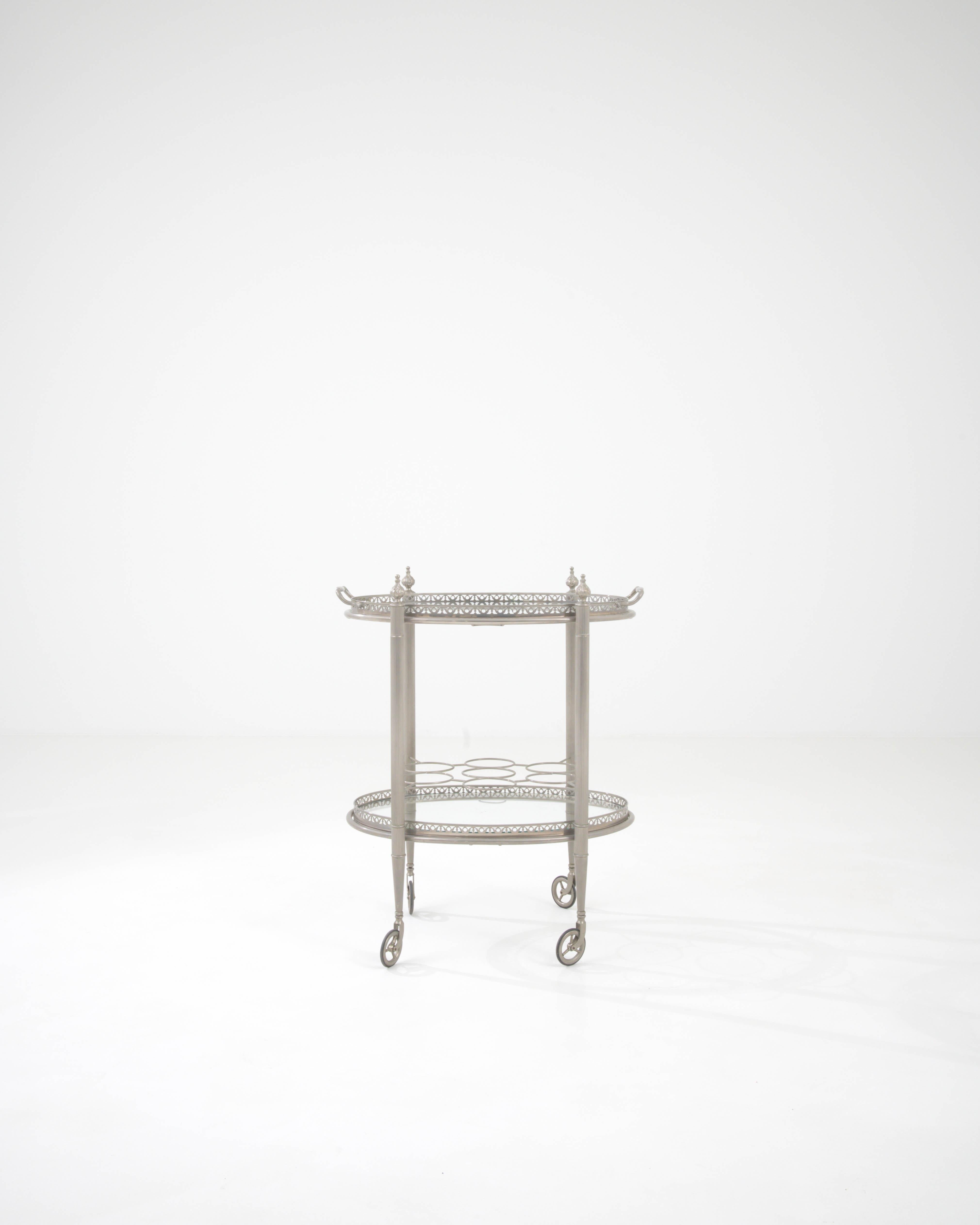This 20th Century French Metal & Glass Bar Cart is a quintessential piece that infuses functional design with a whisper of romantic nostalgia. Fashioned from fine metal, its frame boasts a silver-tone finish, giving it a subtle glow that complements