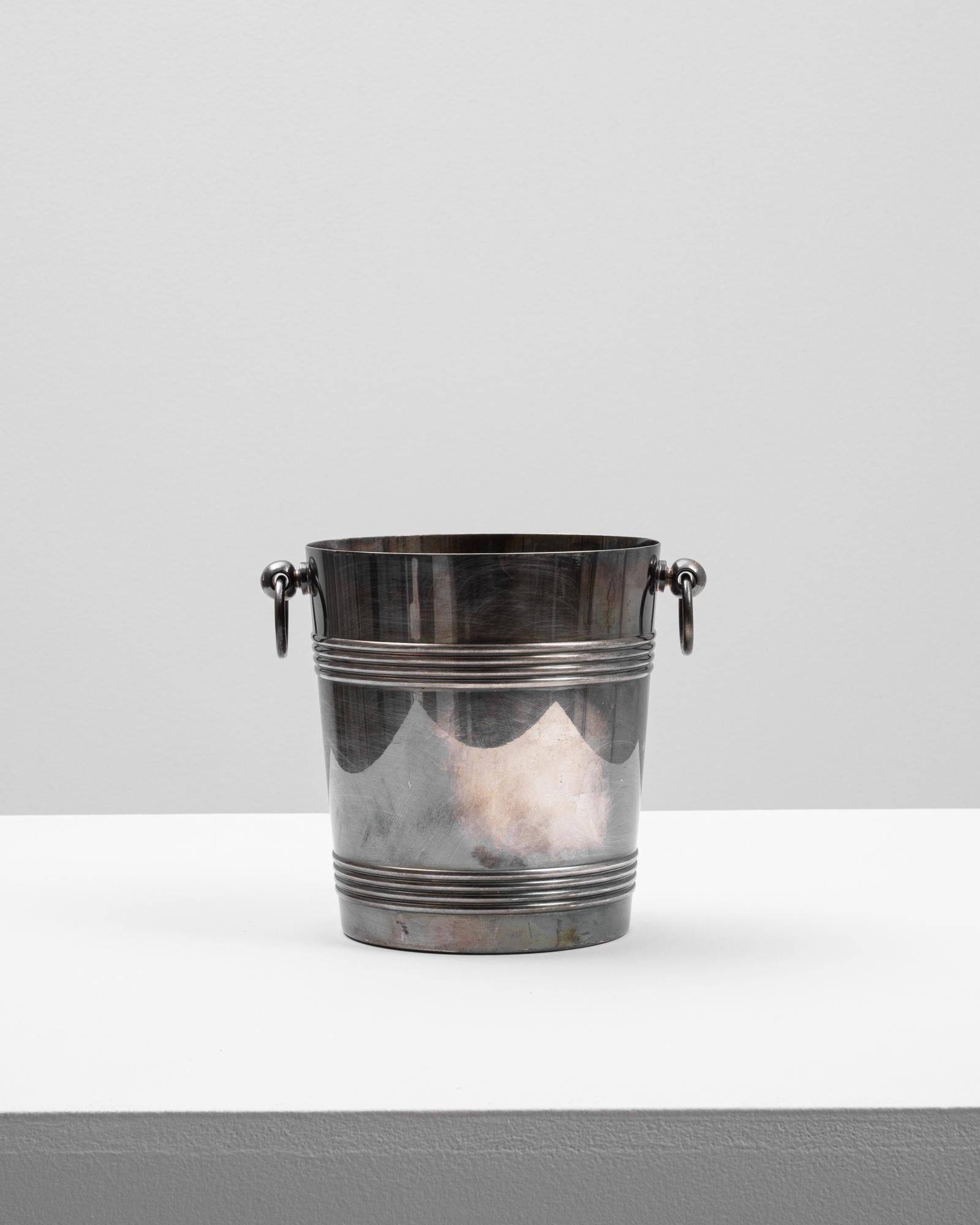 This 20th Century French metal ice bucket combines sleek design with subtle decorative detail. Made from polished metal with a slightly aged patina, the bucket features double banded accents at the top and base, along with a scalloped motif around