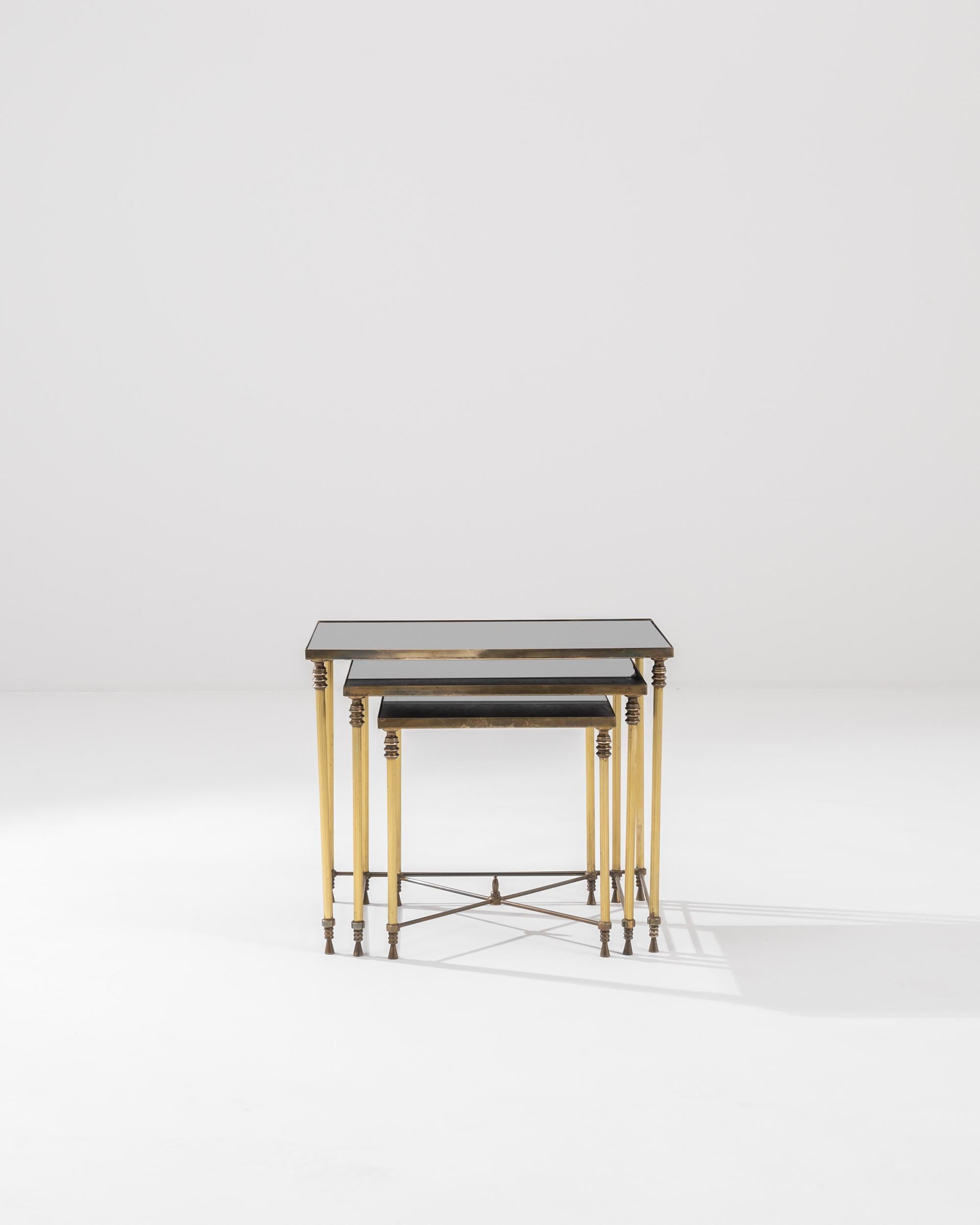 A set of metal nesting tables with glass tops from 20th century, France. Brass base, gold-colored legs, and darkly tinted glass form a pleasingly bright color palette. The patina that has spread across the metal indicates the mature age of these