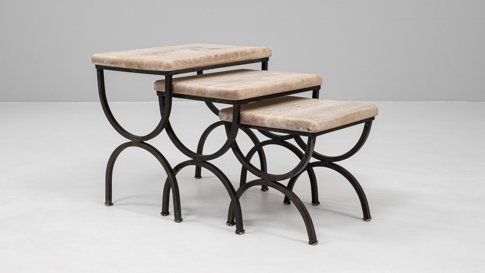 This set of three 20th Century French nesting tables features a striking combination of robust materials and sculptural form. Each table showcases a gracefully arched blackened metal base, paired with a lightly patinated wooden top that brings