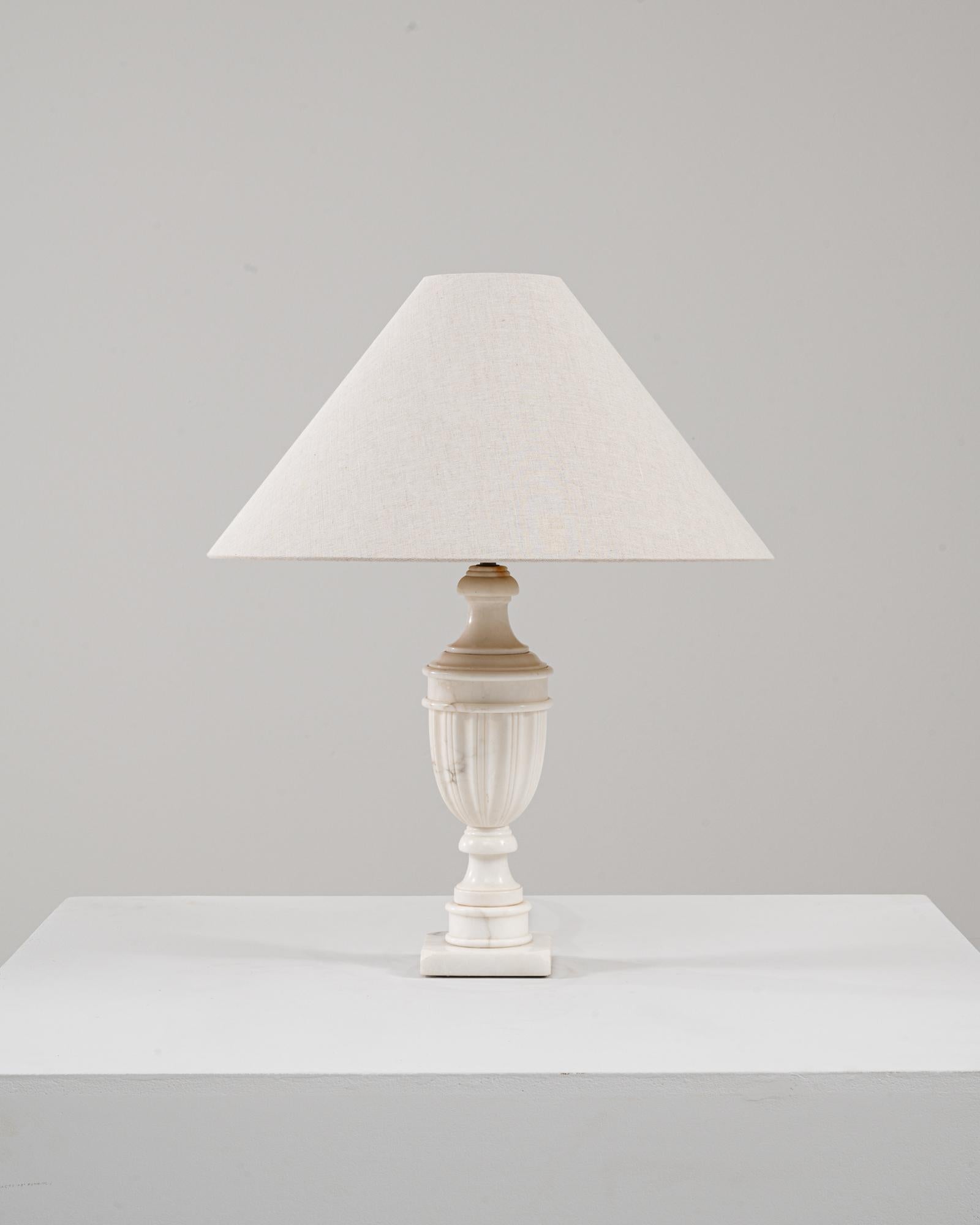 Esta lámpara de sobremesa de mármol neoclásica francesa del siglo XX ofrece una presencia elegante y escultural anclada en el diseño clásico. El cuerpo en forma de urna está tallado en mármol cremoso con suaves vetas, lo que confiere a la pieza una