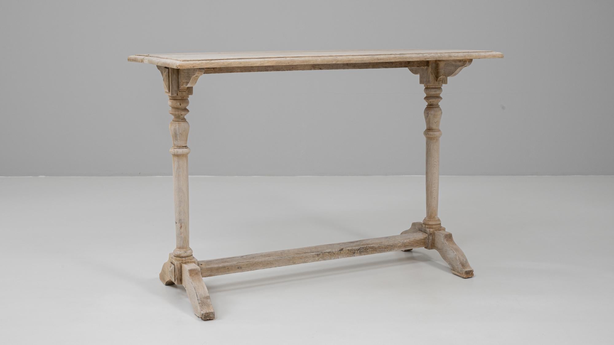 This 20th Century French oak console table showcases a refined rustic aesthetic rooted in traditional craftsmanship. The piece features a long, narrow top with softly worn edges, supported by two elegantly turned legs that introduce subtle classical