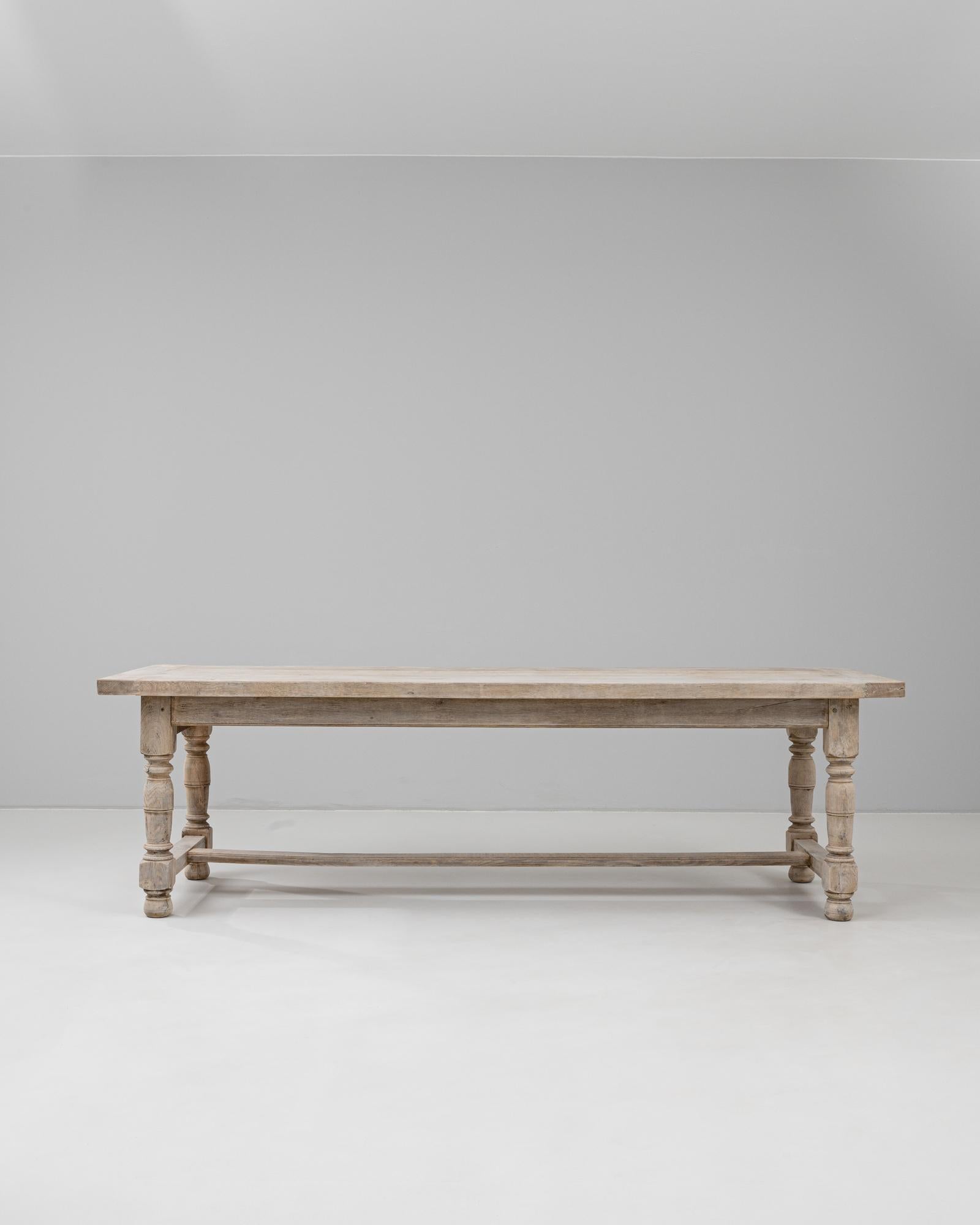 This 20th Century French dining table is crafted from light oak, showcasing a pale, matte finish that enhances its natural grain and understated elegance. The rectangular top rests on four turned legs connected by a stretcher, offering both