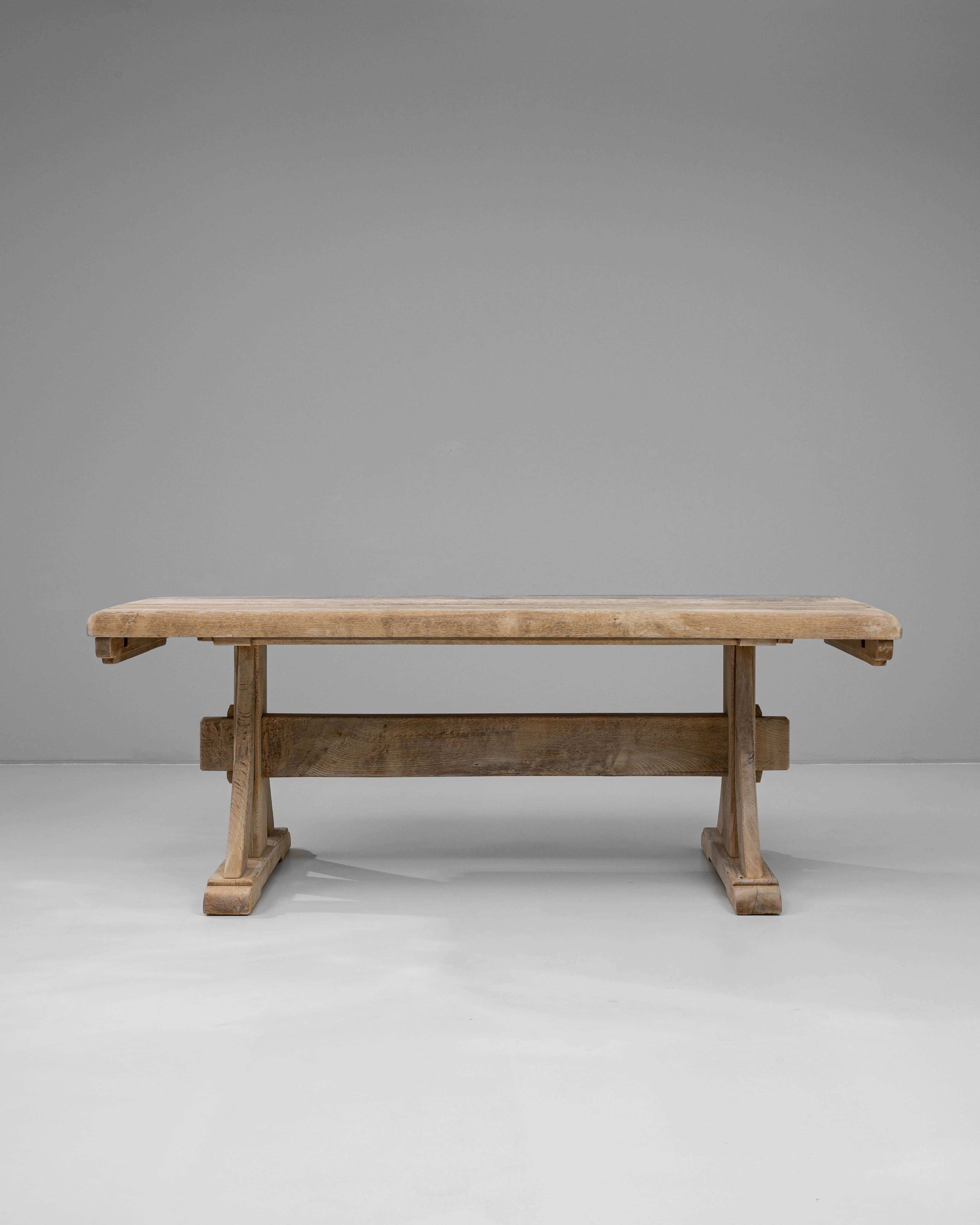 This 20th-century French oak dining table features a bold, architectural form that highlights the beauty and strength of solid oak craftsmanship. Its rectangular top rests on a sturdy trestle base with carved supports and a central stretcher,