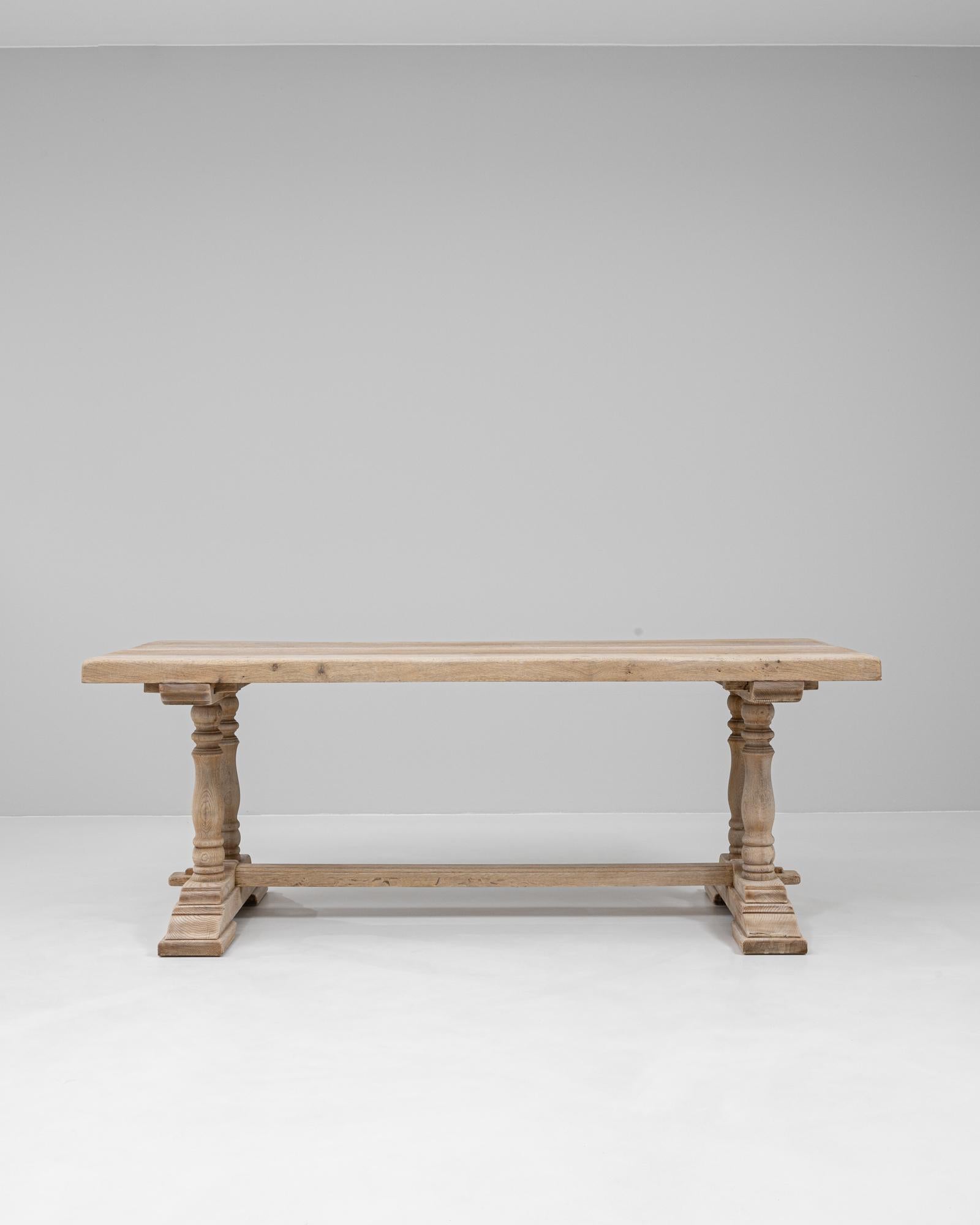 This 20th century French oak dining table showcases a refined balance of craftsmanship and rustic charm. The solid oak construction features a rectangular plank top supported by double baluster legs connected by a central stretcher for added