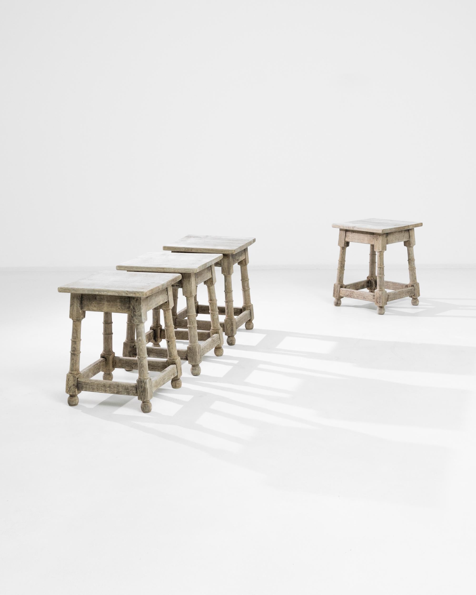 A set of four 20th century wooden stools produced in France. Four bleached oak stools featuring splayed legs and bun feet, joined by square stretchers and assembled with mortise and tenon joints. The carved legs exhibit four turned rings and