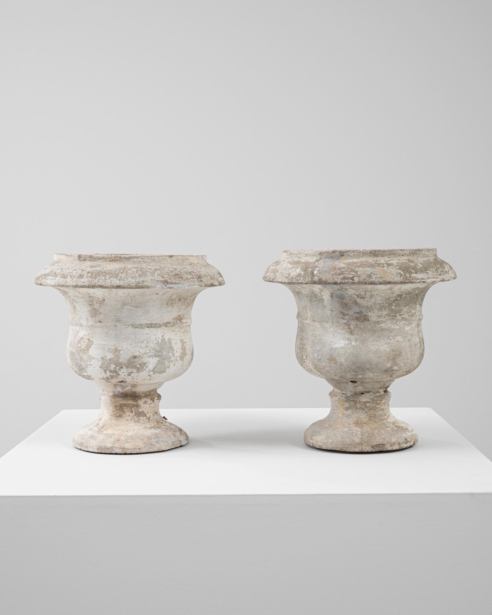 This pair of 20th Century French concrete planters features a classic urn silhouette with a flared rim, rounded body, and pedestal base. Crafted from durable cast concrete, each piece exhibits a softly weathered surface with traces of mossy patina