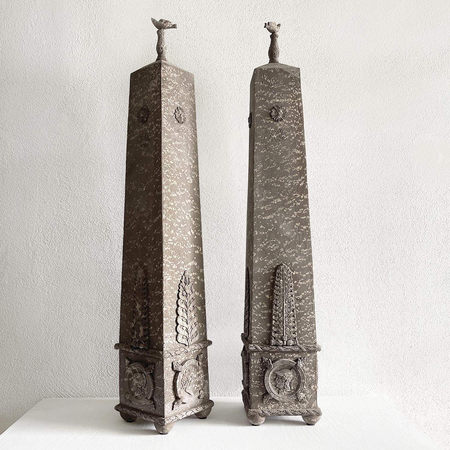 A vintage pair of terre cuite Obelisks in the new classical style, in good condition. The lower part is adorned with small reliefs of mythological profiles - the bodies are decorated with large acanthus leaves, and topped with a small bird. Wear