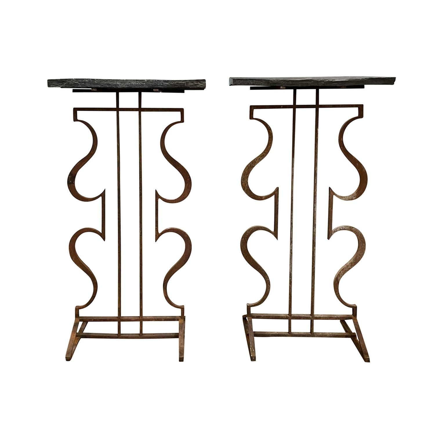 A vintage French pair of rustic console tables featuring heavy slate tops with rough edges, in good condition. The freestanding end tables are supported by elegantly curved hand-forged iron bases. Suitable for both indoor and outdoor use, these