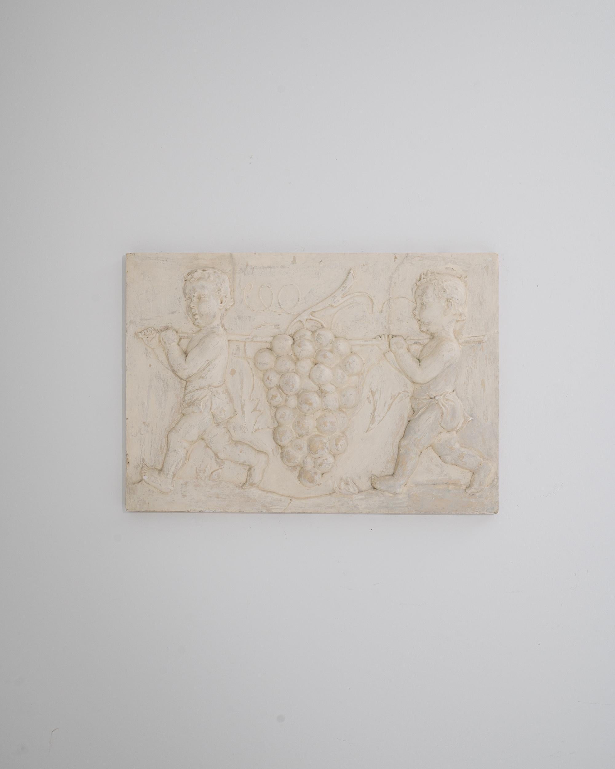 A vintage plaster wall decoration depicting two putti carrying an enormous bunch of grapes. Made in France in the 20th century, the white plaster relief evokes the decorative arts of the French academy. The playful game of scale between the size of