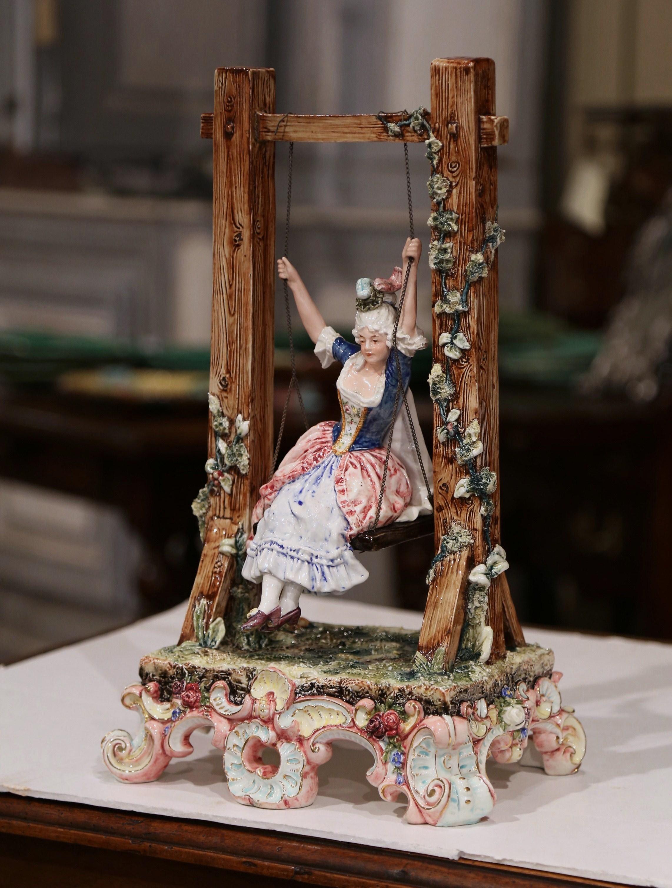 Decorate a shelf or a bookcase with this colorful antique Majolica figure sculpture. Crafted in France, circa 1920, the large, intricate, sculptural piece features a young woman balancing on a swing in a courtly costume. The style and scene are