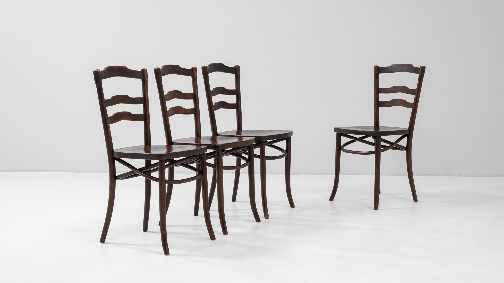 This 20th century French set of four dining chairs offers a classic country aesthetic, featuring gently curved ladder backs and rounded wooden seats. Crafted from dark-stained wood, each chair showcases a refined rusticity with its elegantly arched
