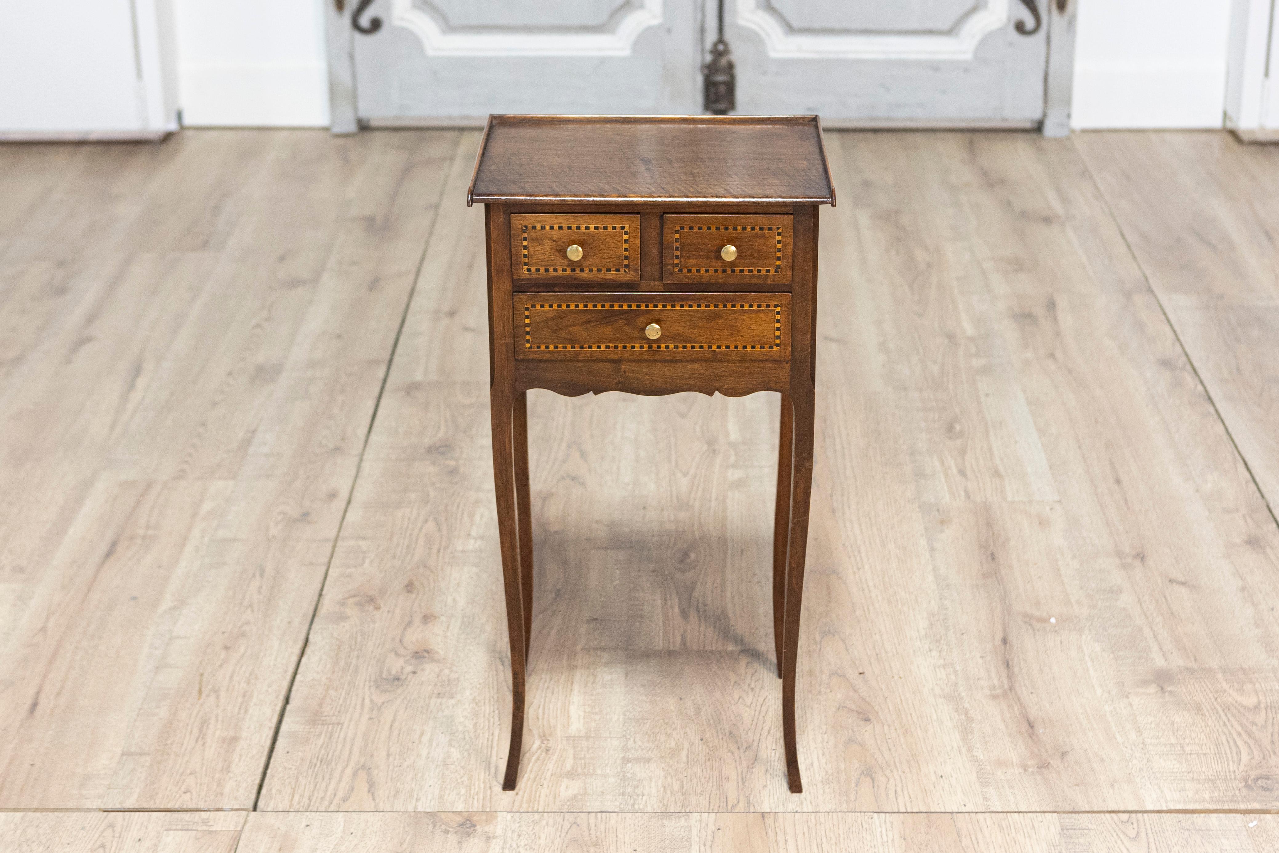 Cette charmante et élégante table d'appoint du début du XXe siècle est fabriquée en noyer massif et présente un grain chaud et riche. Il comporte trois tiroirs en façade disposés sur deux rangs, chacun orné de délicates incrustations de filets de
