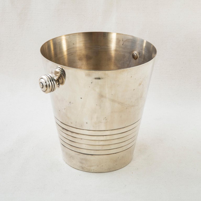 This French vintage silver plate champagne bucket features concentric circles around the top and bottom. Two knob handles allow for easier carrying. The bottom is stamped Auchan, 25 ans, 26-10-86 commemorating the 25th anniversary of the company.