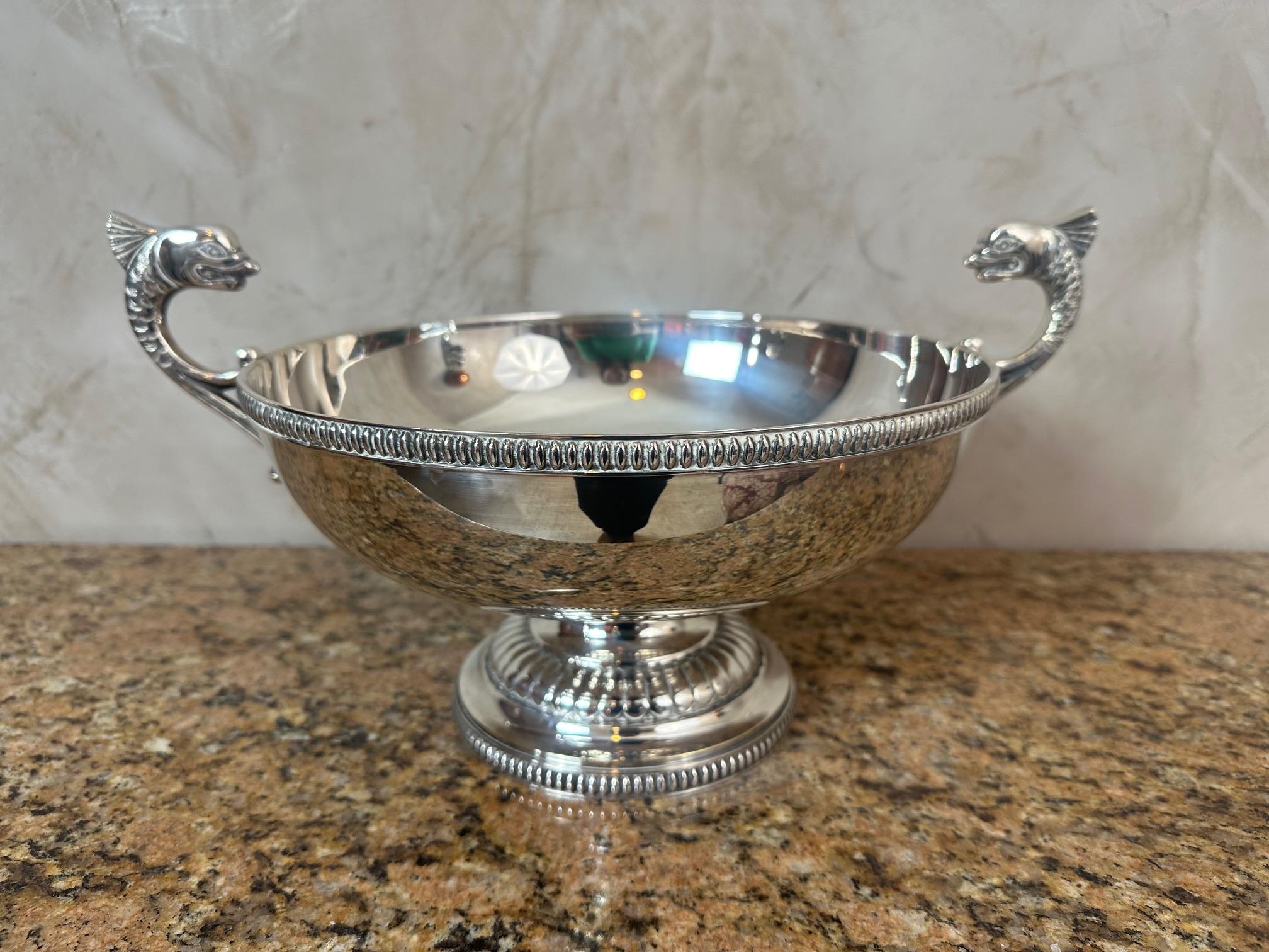 Élégante coupe à fruits en métal argenté datant des années 1920, reposant sur une base piédestale moulée avec une délicate bordure perlée. La large coupe circulaire est ornée de deux anses en forme de dauphin finement détaillées, ajoutant à la fois