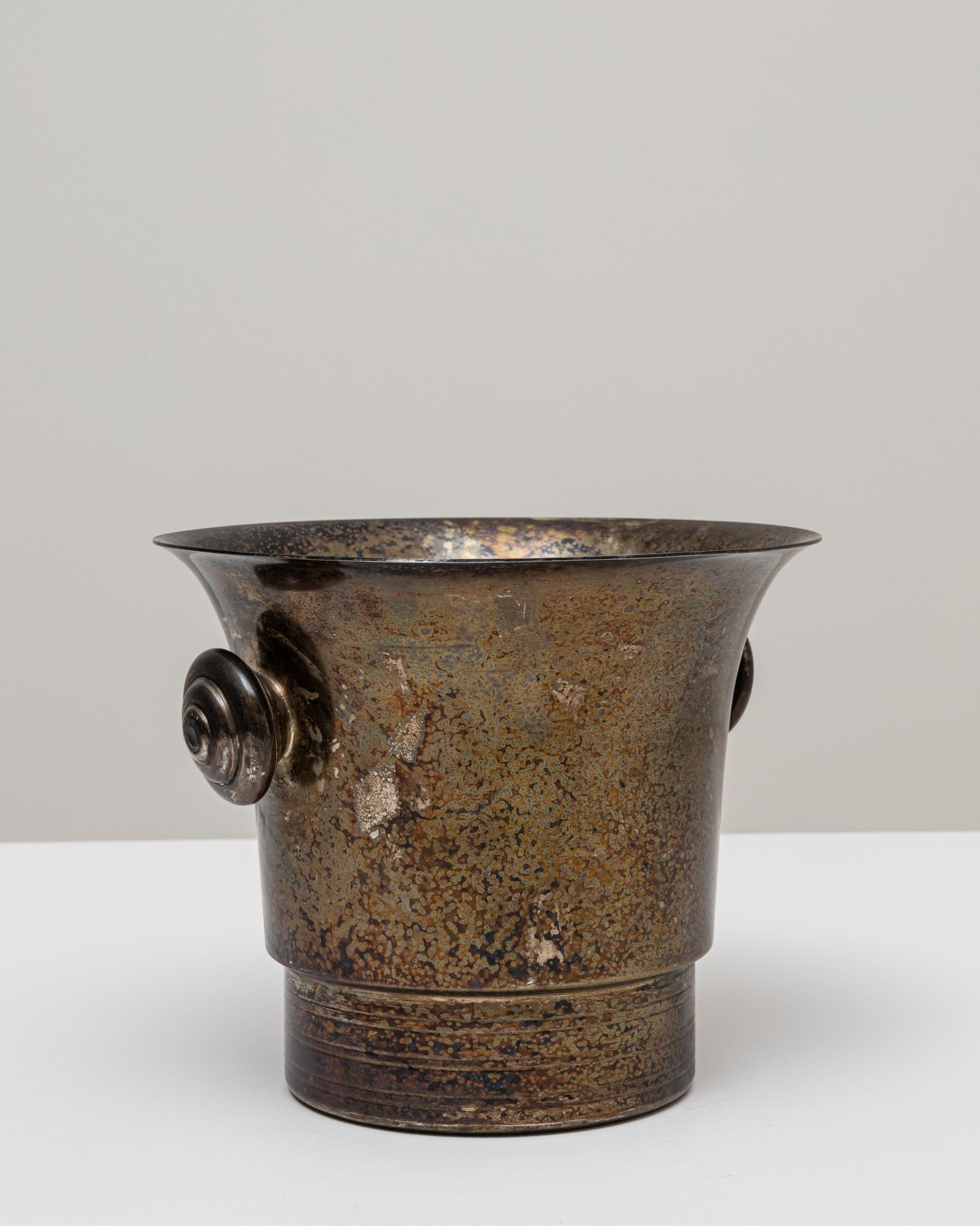 This 20th Century French silver-plated champagne bucket presents a beautifully aged surface, marked by a rich, mottled patina that enhances its character and sense of history. Its flared rim, cylindrical base, and sculpted side handles give the