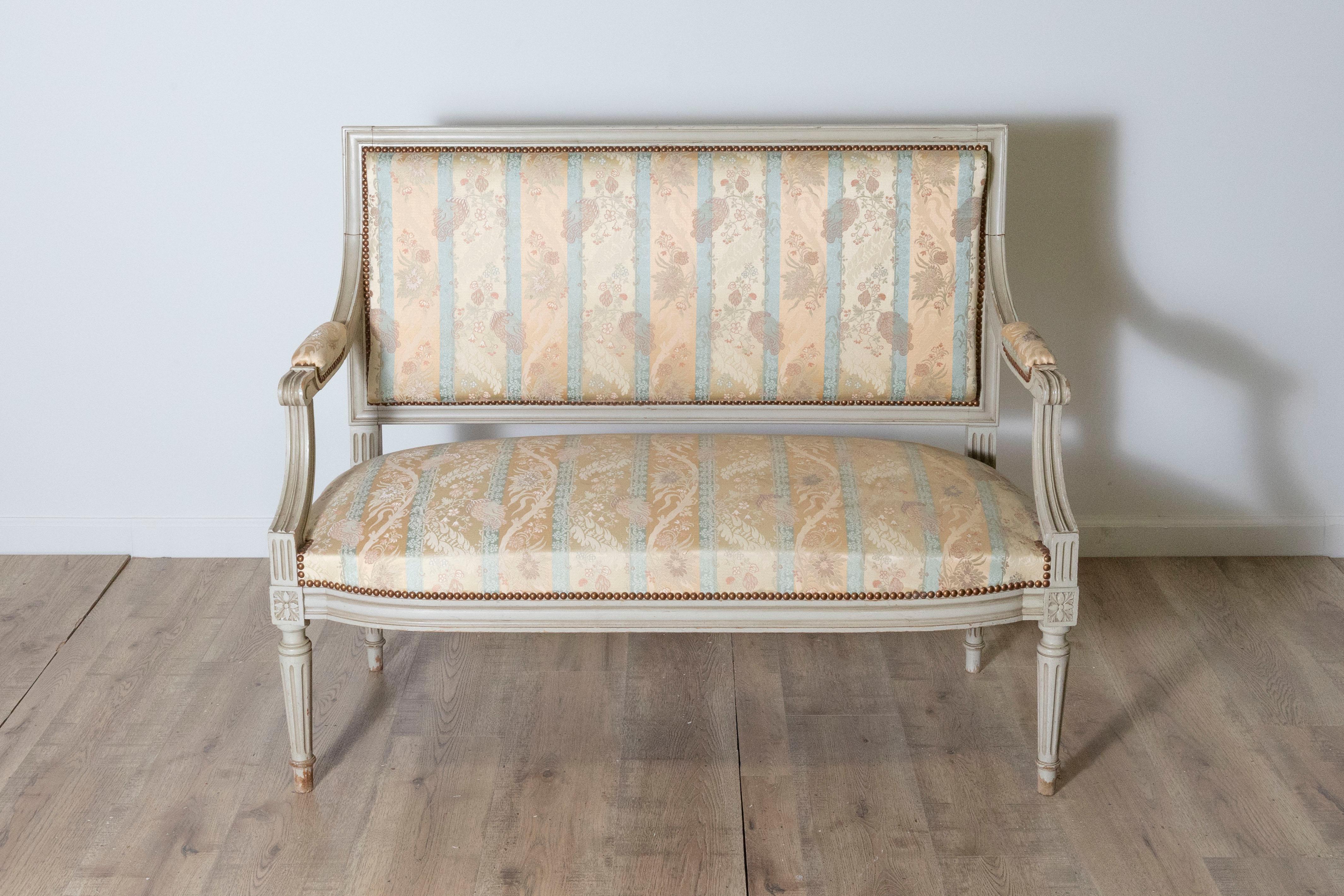Questo elegante divano francese del XX secolo esemplifica lo stile Luigi XVI, combinando elementi di design neoclassico con una raffinatezza senza tempo. La struttura in legno massiccio di faggio è intagliata a mano con gambe scanalate, accenti di