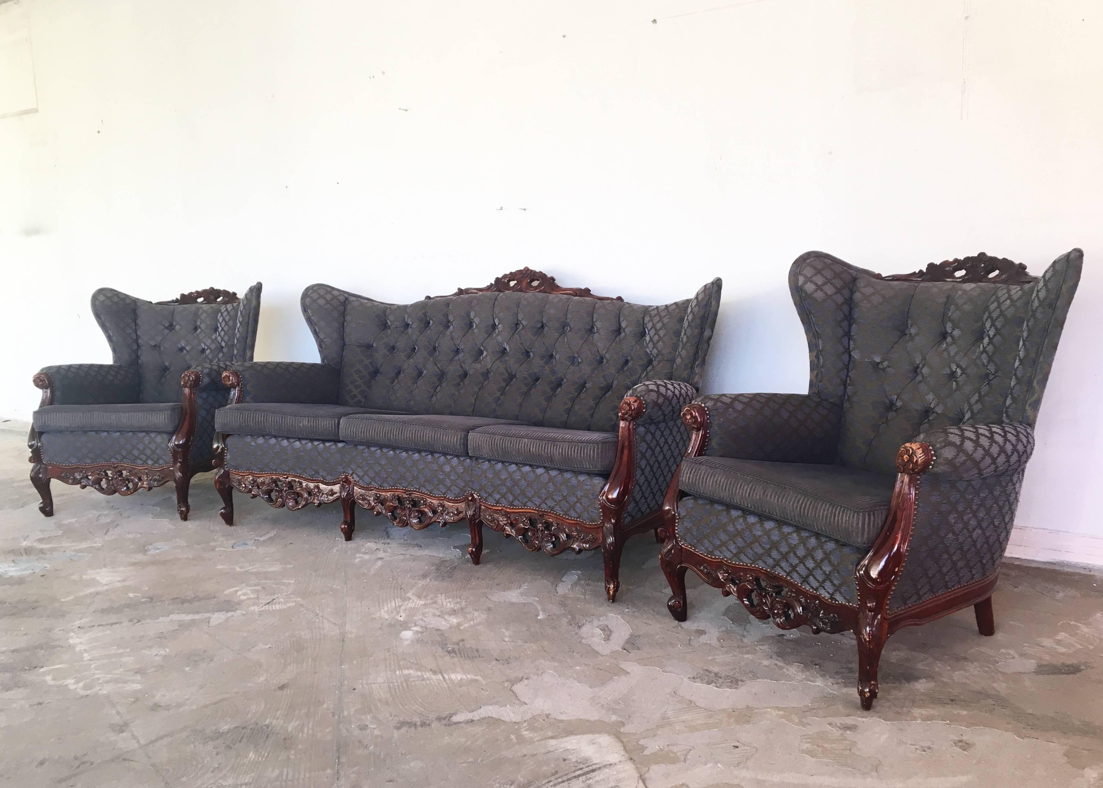 This three-piece living room or sofa set, consists of a three-seat sofa and two wingback armchairs. The set features sculptural carved wood and capititonné backrests. The fabric overall looks purple, blue or gray with gold, depending how the light