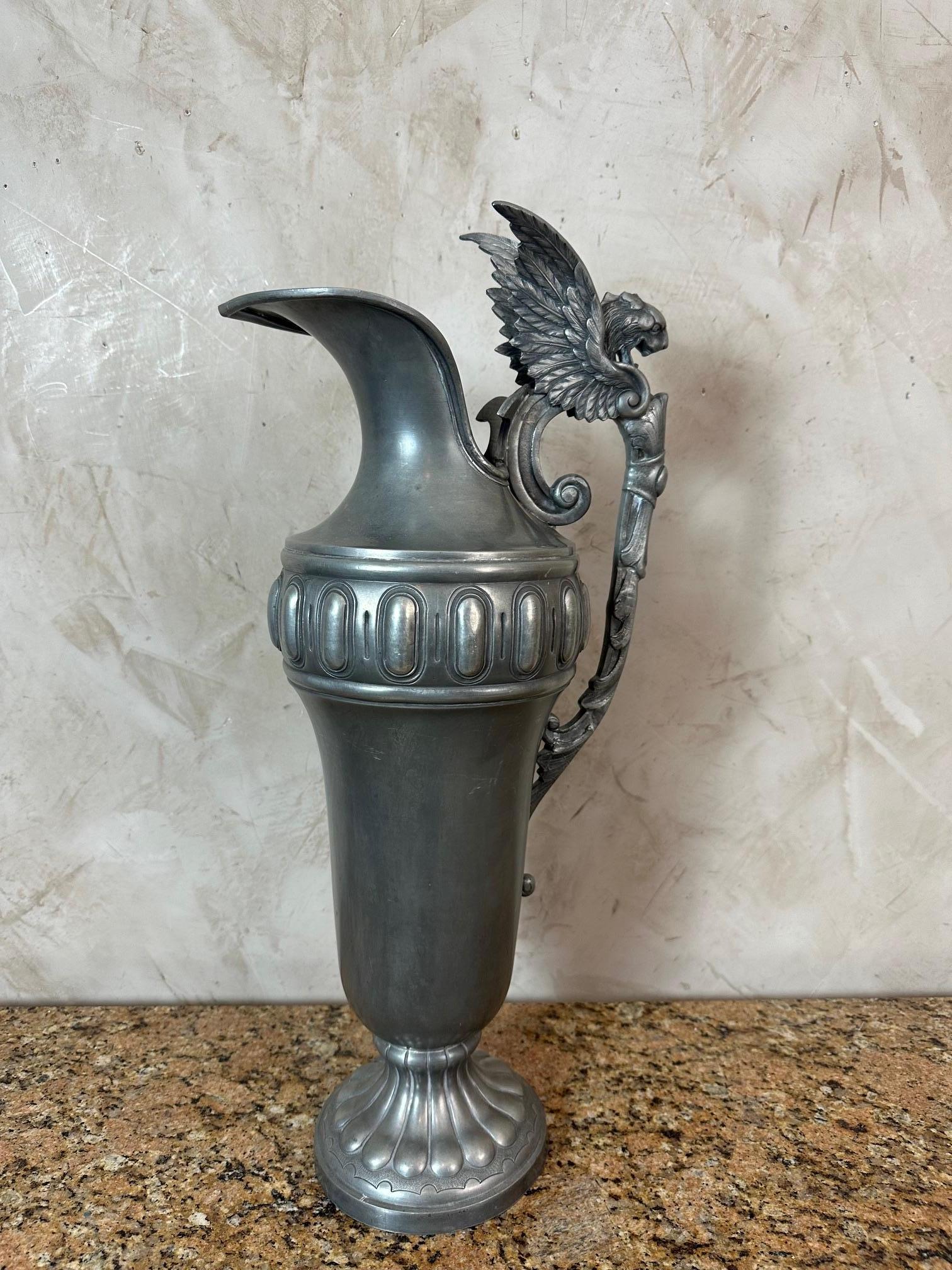 Aiguière en étain raffinée datant des années 1930, présentant une belle patine et un design finement détaillé inspiré du style néoclassique.
Sa poignée, délicatement sculptée en forme d'aile et de tête de griffon, confère à la pièce un caractère