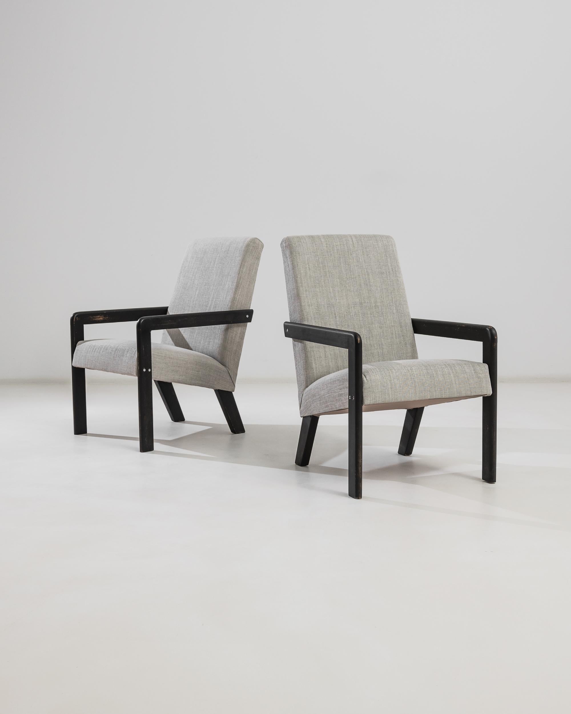 Add a touch of mid-century modern charm to your home with this pair of 20th Century French Upholstered Armchairs. These stylish chairs feature sleek black wooden frames that provide a striking contrast to the soft, grey upholstery. The clean lines