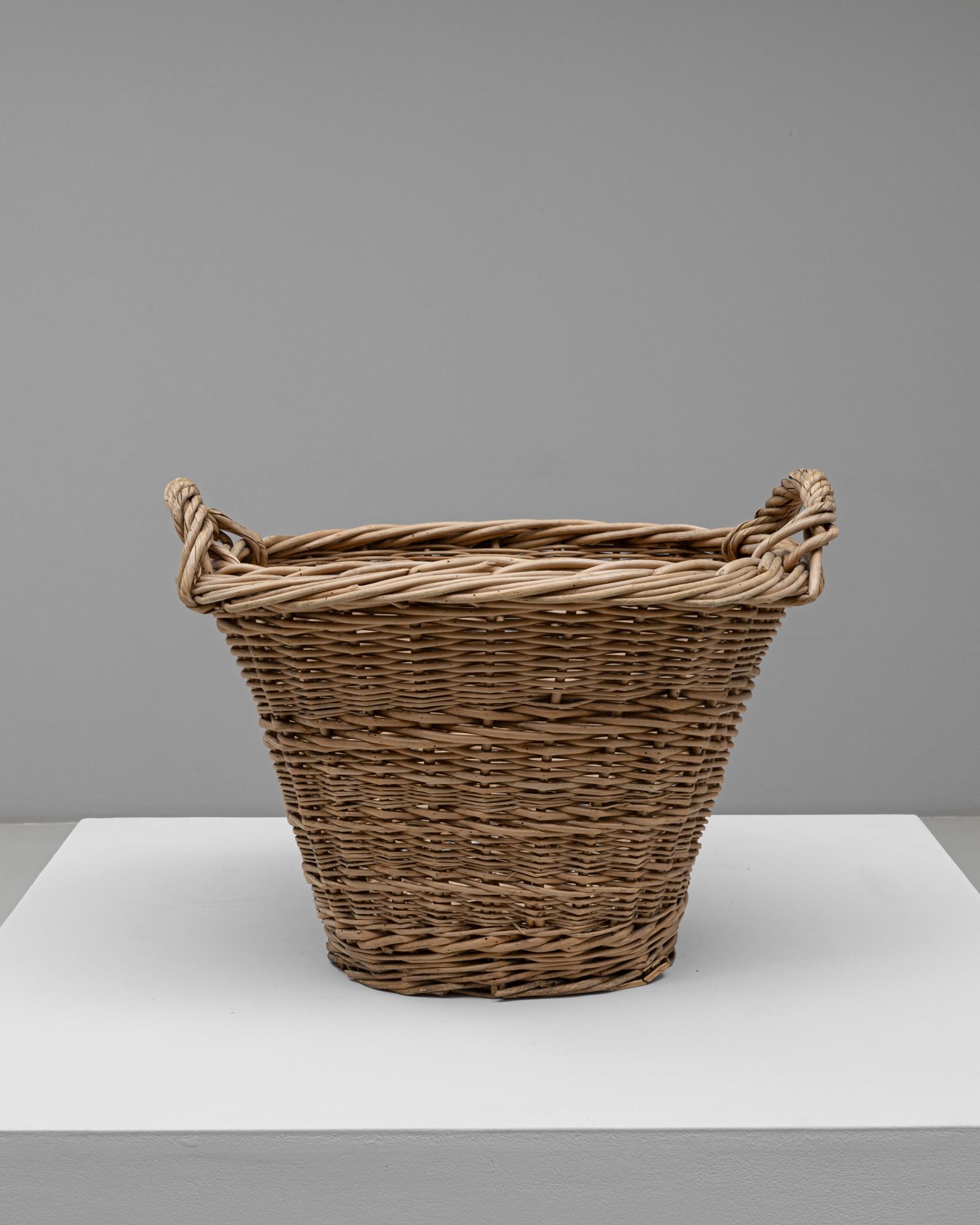 This 20th Century French wicker basket showcases traditional handwoven craftsmanship and timeless rustic appeal. Made from natural wicker, it features a wide, tapered form with thick, twisted rope handles on each side for durability and ease of