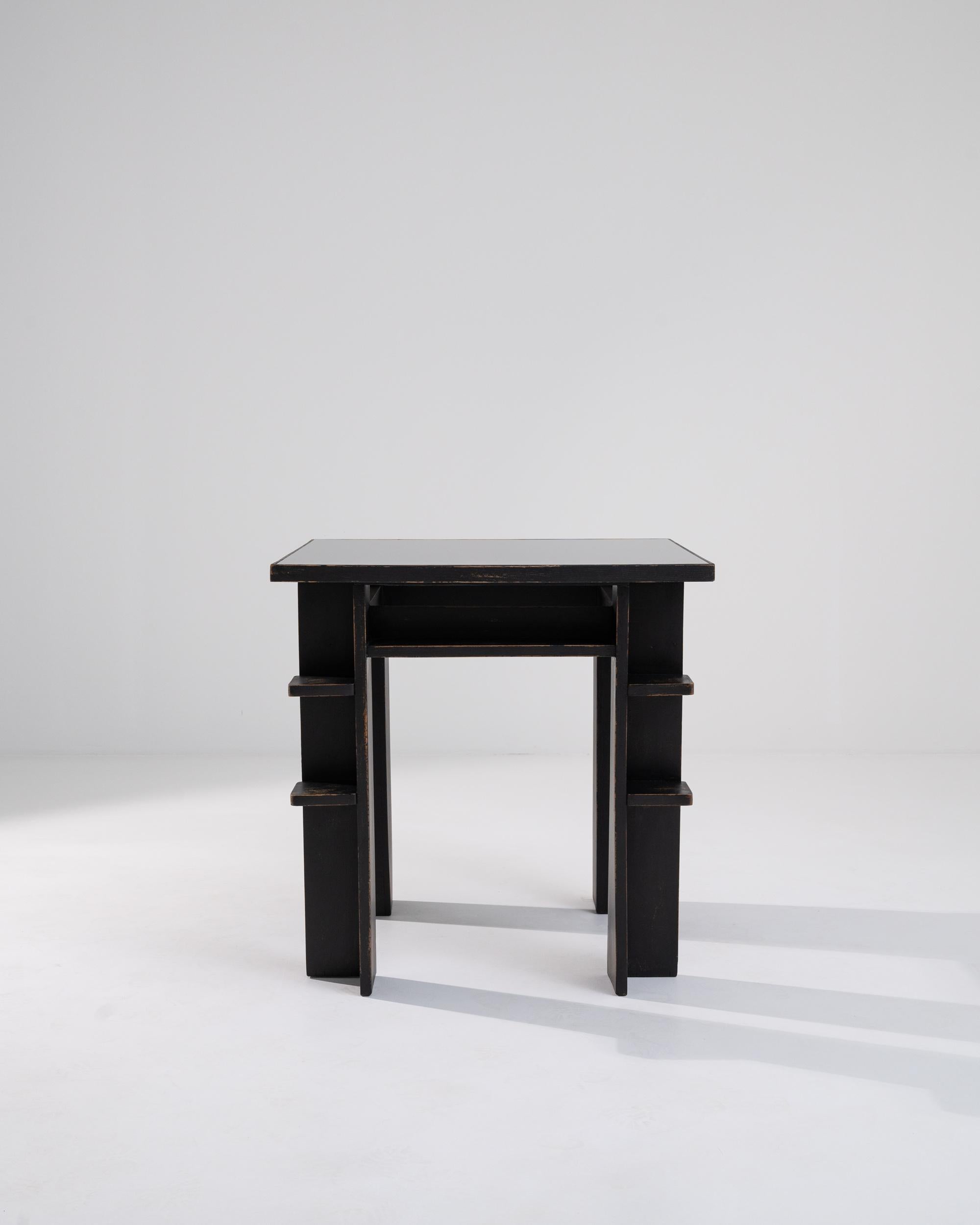 A wooden table with a glass top created in 20th century France. This minimal side table is designed in an early Modern style, constructed by right-angled legs slotted with curious little shelves. A rich coat of black paint has been slowly worn away