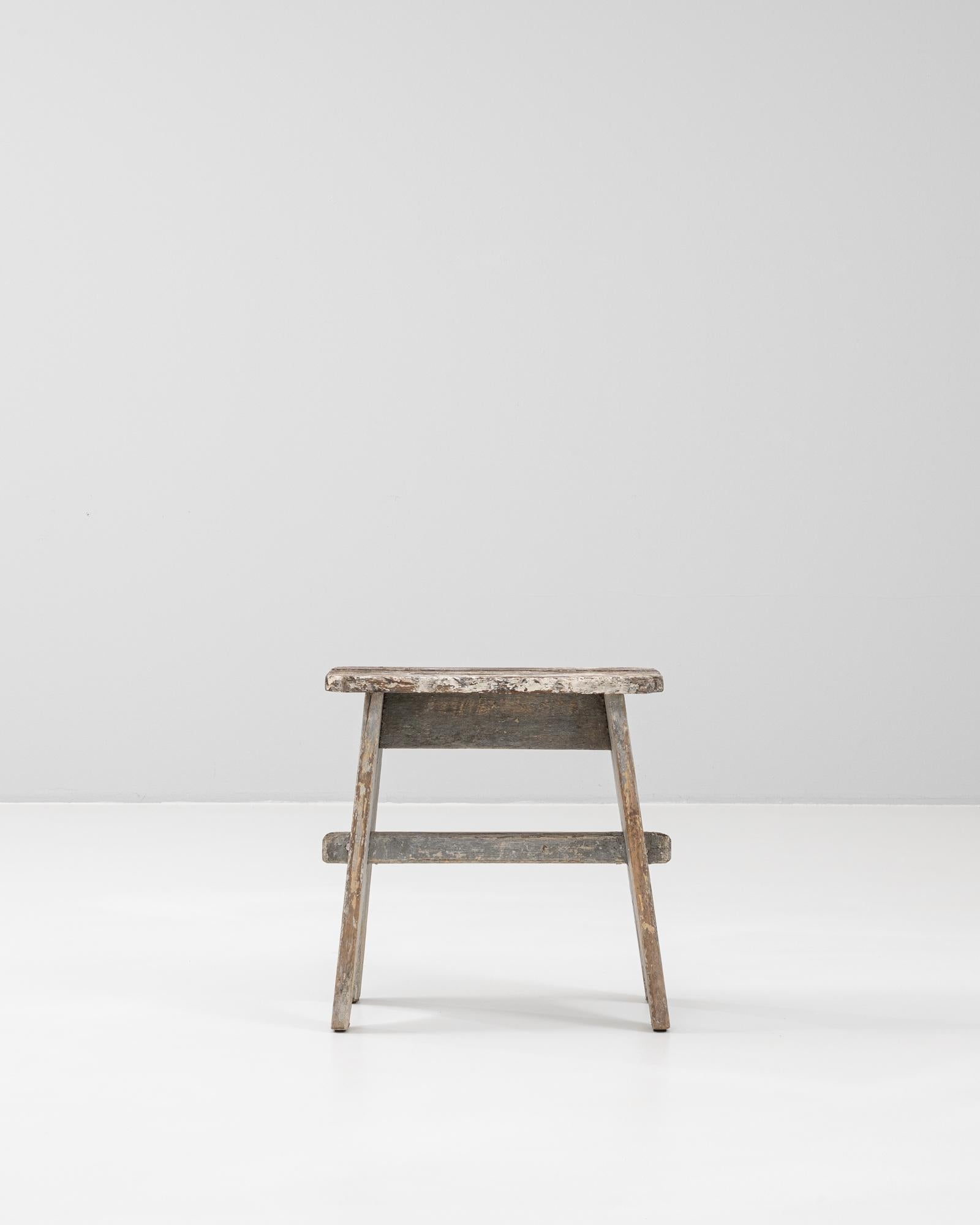 This 20th Century French wooden stool exudes rustic charm with its weathered patina and timeworn character. Crafted from solid wood, the stool features a simple, functional form with splayed legs joined by stretchers and a thick, rectangular seat.