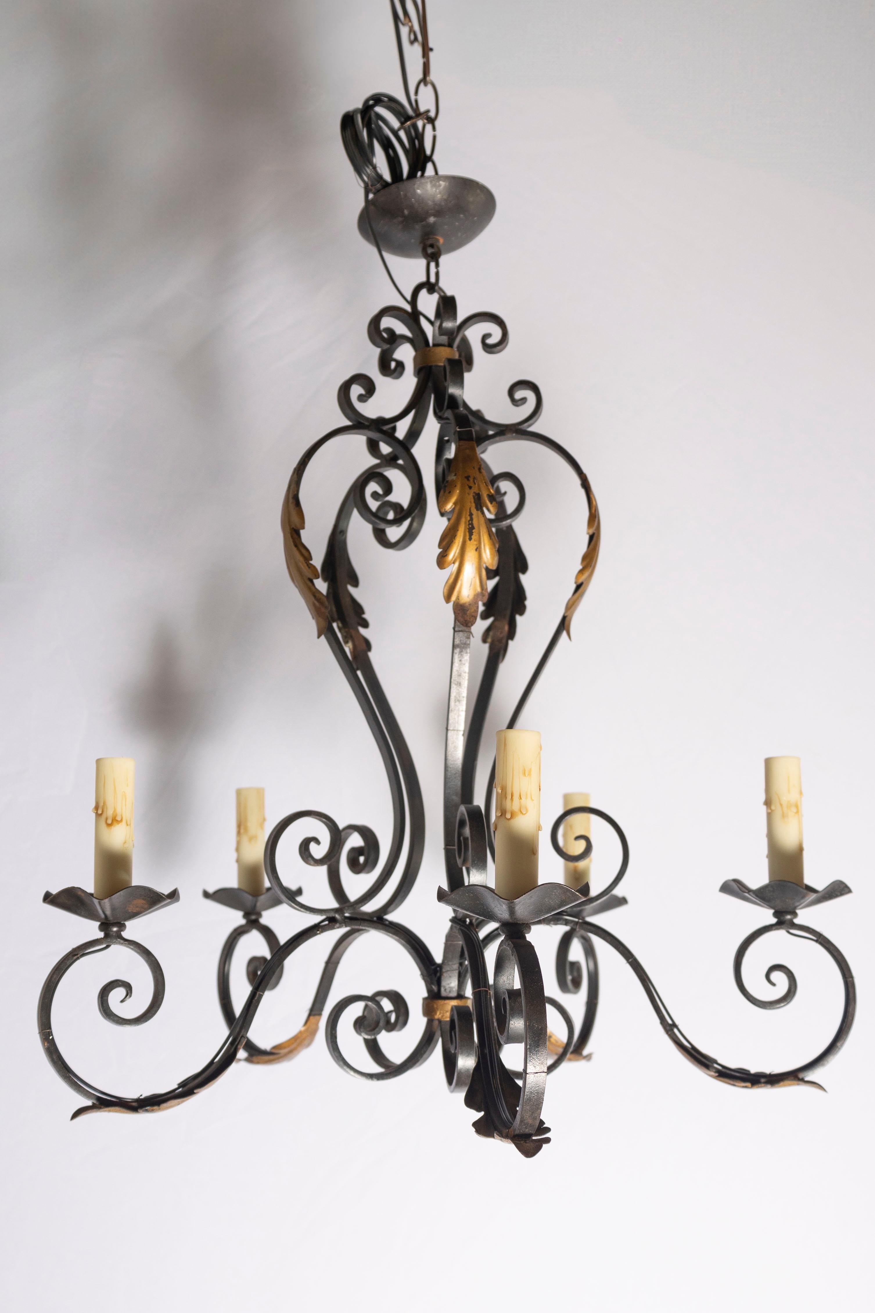 Questo elegante lampadario francese della metà del XX secolo è una straordinaria espressione della lavorazione artigianale del ferro, che unisce la forza rustica alla raffinatezza ornamentale. Realizzato a mano in ferro battuto, il lampadario
