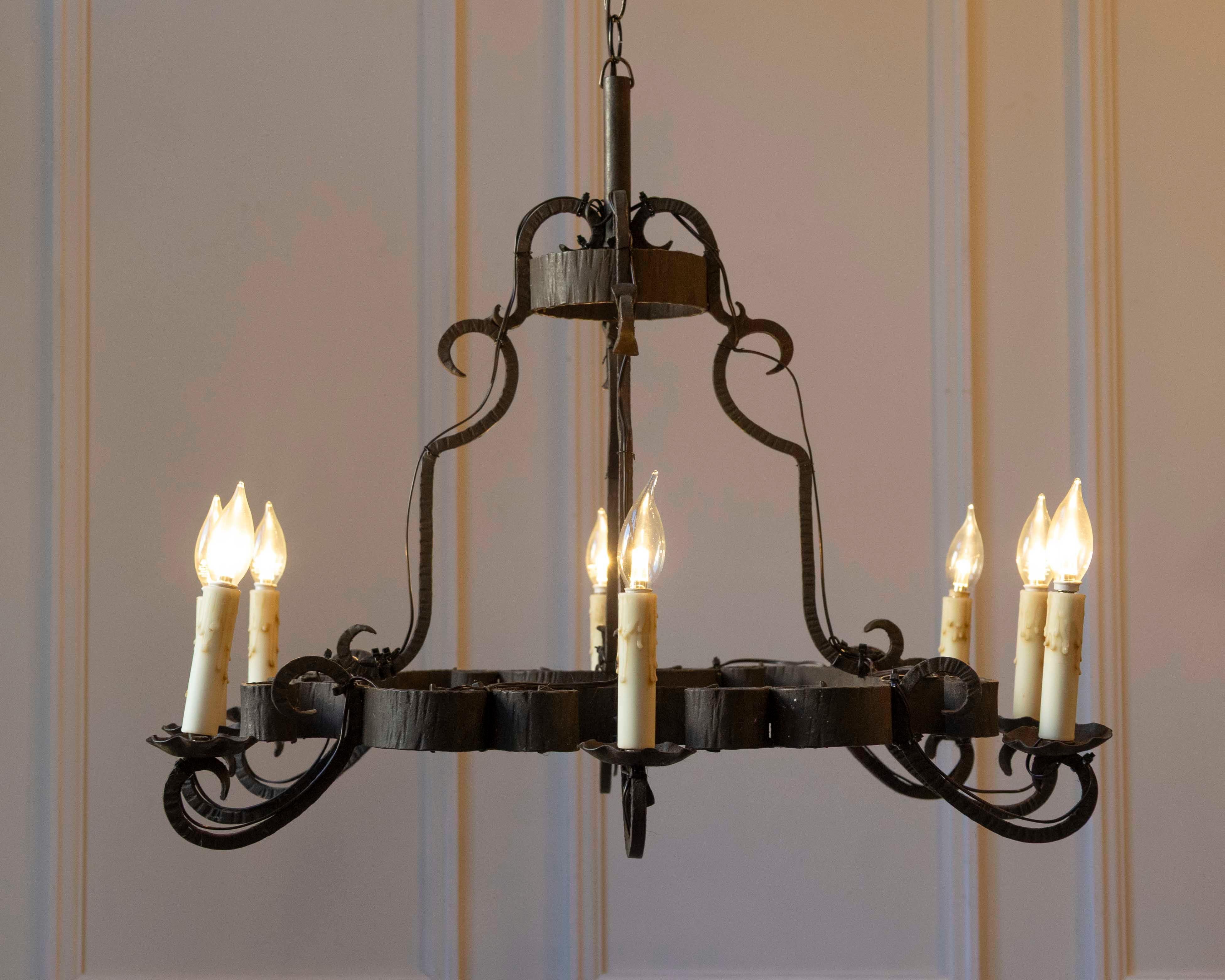 This 20th-century French wrought iron chandelier presents a substantial, hand-forged design built around a circular iron ring. Eight scroll-form arms extend outward in a consistent rhythm, each supporting candle-style lights with shaped bobeches.