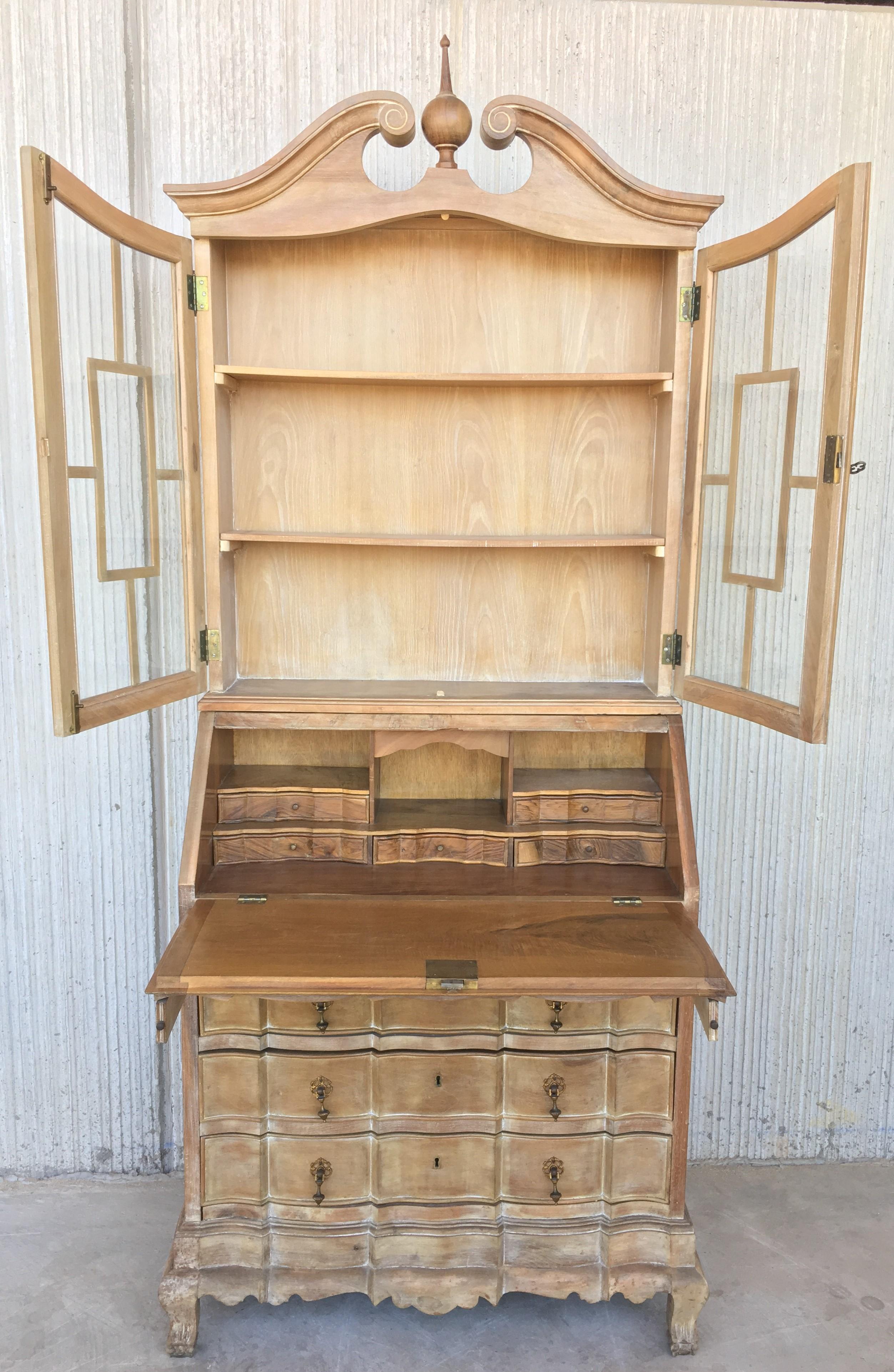 With a broken bonnet with rosette form ends and an urn form finial. The shaped burled wood double glass door opens to two shelves. The slant front opens to cubby holes, five drawers. All with brass bat wing hardware and escutcheons. Raised on
