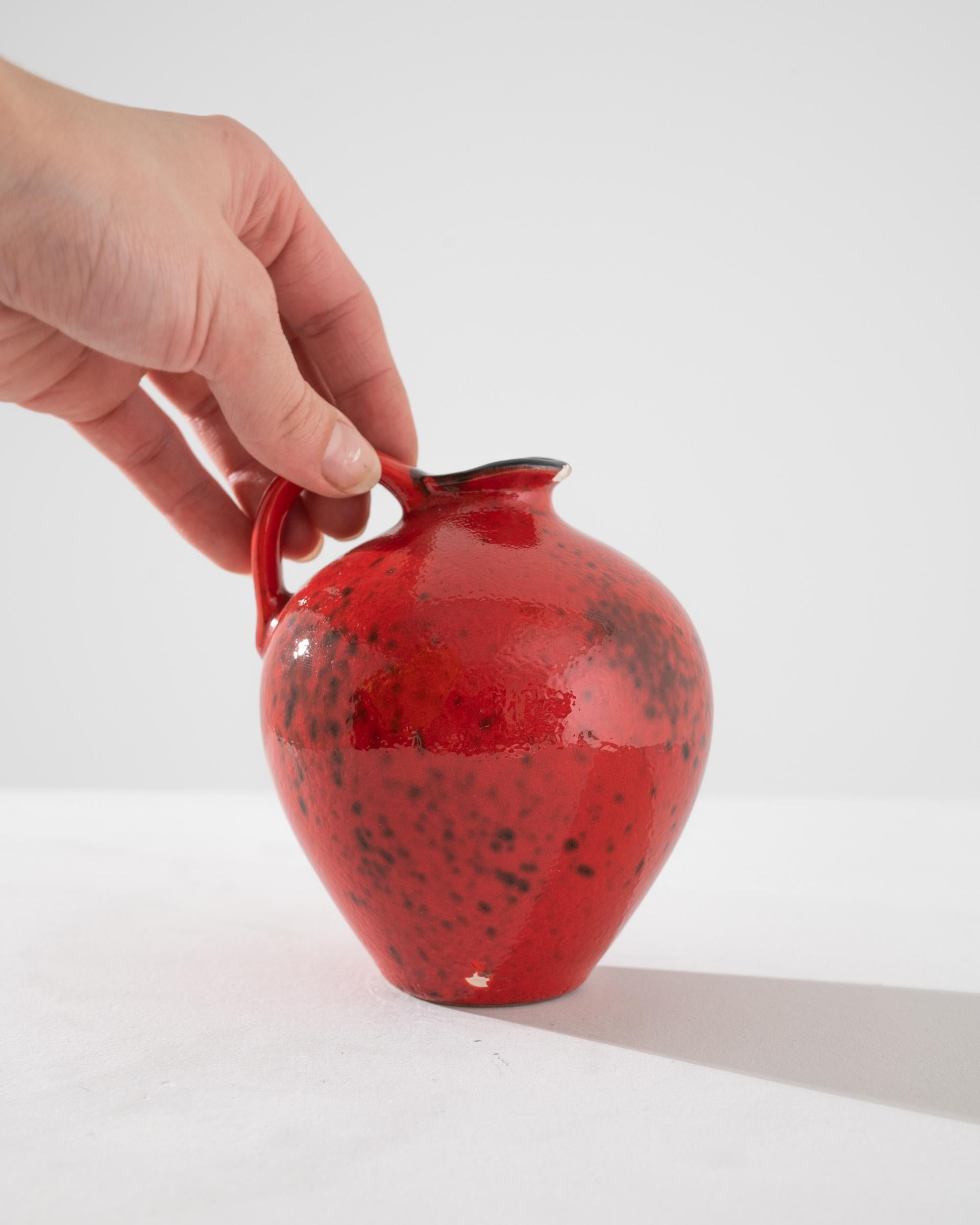 Cette cruche en céramique allemande du XXe siècle est un exemple frappant d'art fonctionnel. Sa glaçure rouge vif, mouchetée d'accents plus sombres, lui confère un aspect volcanique vivant qui attire l'attention tout en étant élégant. La forme