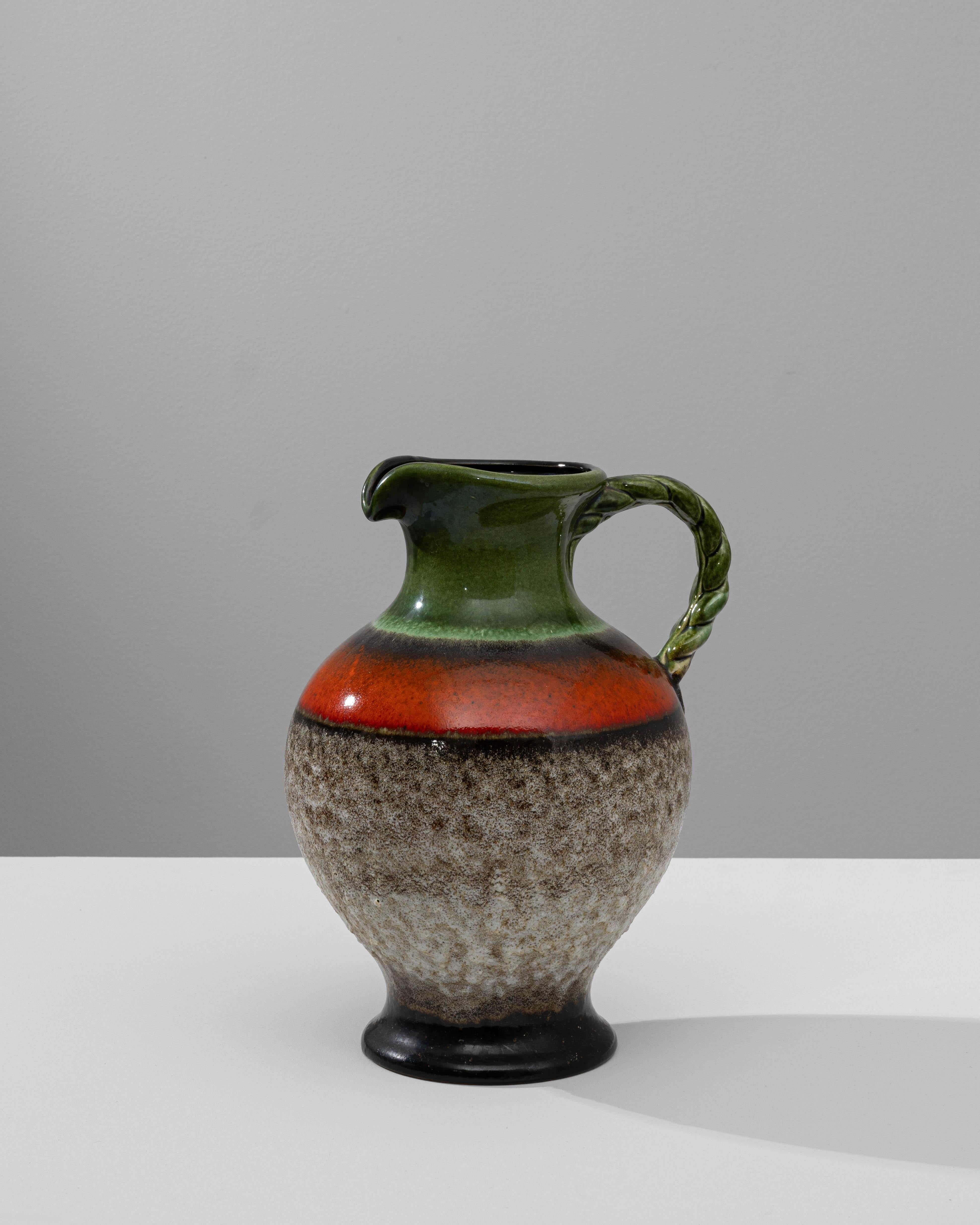 This 20th-century German ceramic pitcher is a stunning example of mid-century artistry, blending vibrant colors and textures. The pitcher features a bold combination of earthy tones, including deep greens and fiery reds, contrasted by a textured,