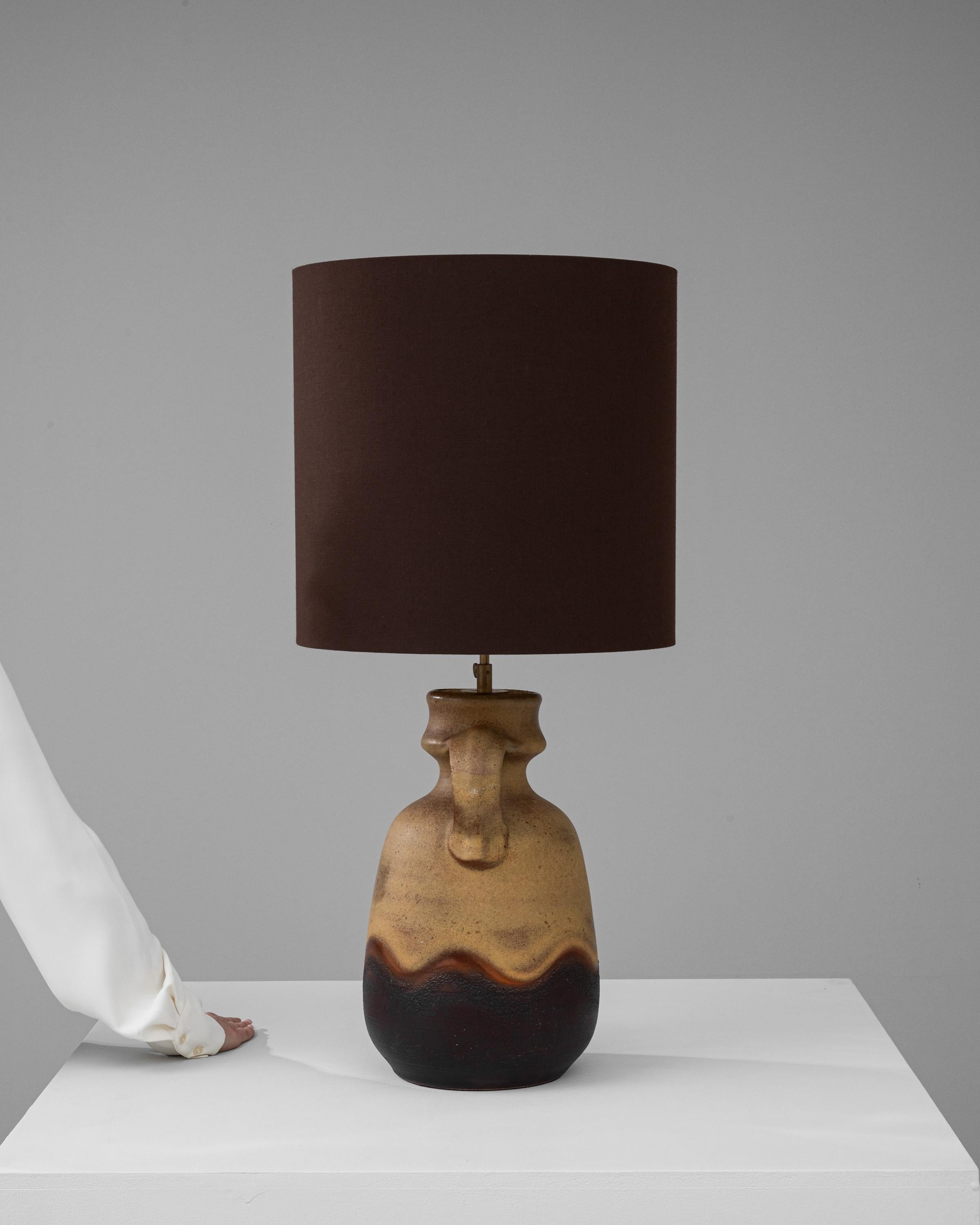 This 20th Century German Ceramic Table Lamp exudes a rustic charm that harkens back to a simpler time. The stout, earthenware base boasts a distinctive handle-like protrusion, a homage to traditional pottery forms, while a rich, earth-toned glaze