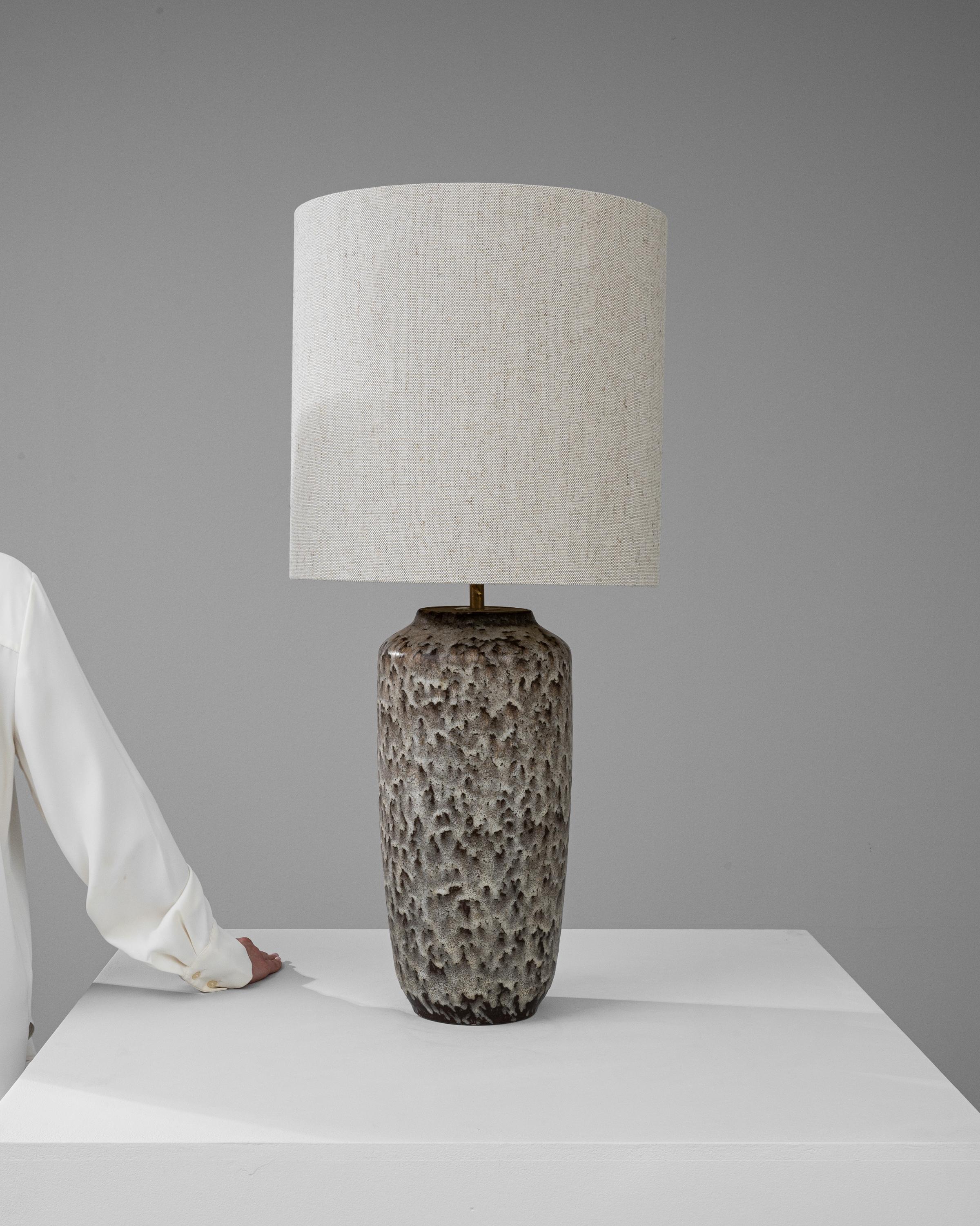 This 20th Century German Ceramic Table Lamp is a masterpiece of texture and form, embodying a rugged natural beauty. The cylindrical base draws the eye with its pitted surface, akin to the surface of a timeworn stone, creating an intriguing tactile