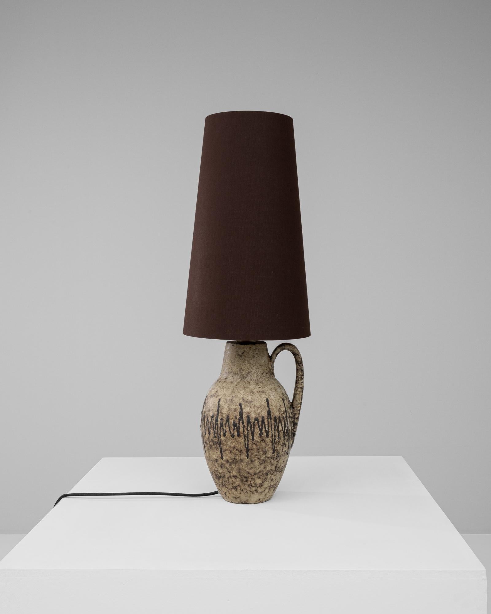 This 20th Century German Ceramic Table Lamp embodies a rustic yet enchanting aesthetic, perfect for those who appreciate the allure of time-honored craftsmanship. The lamp's base, shaped like a classic earthenware jug, features a raw, sandy texture