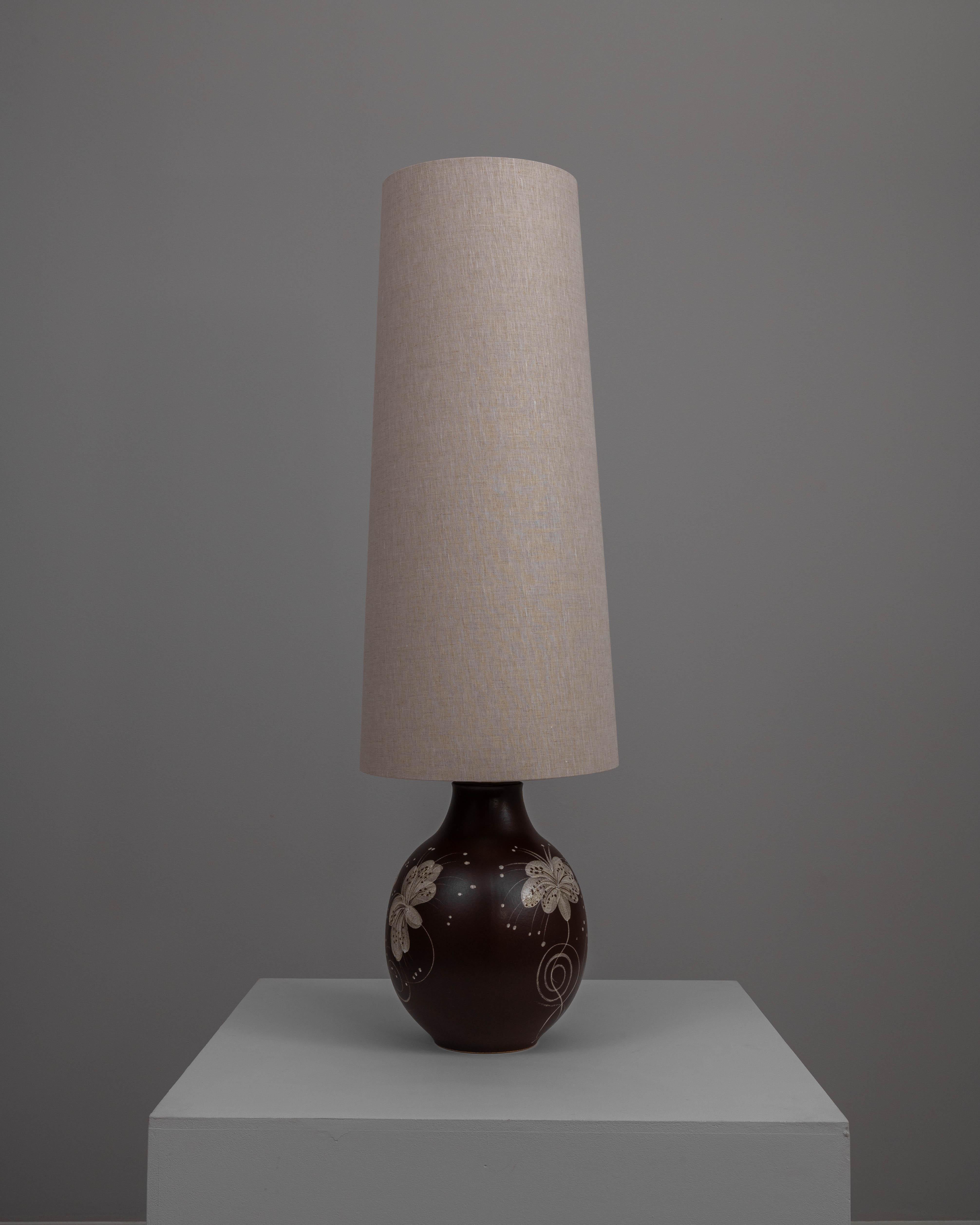 This 20th Century German Ceramic Table Lamp is an exquisite example of functional art. The ceramic base, rich in a deep brown hue, is adorned with delicate white floral motifs that add a touch of elegance and visual interest. These hand-painted