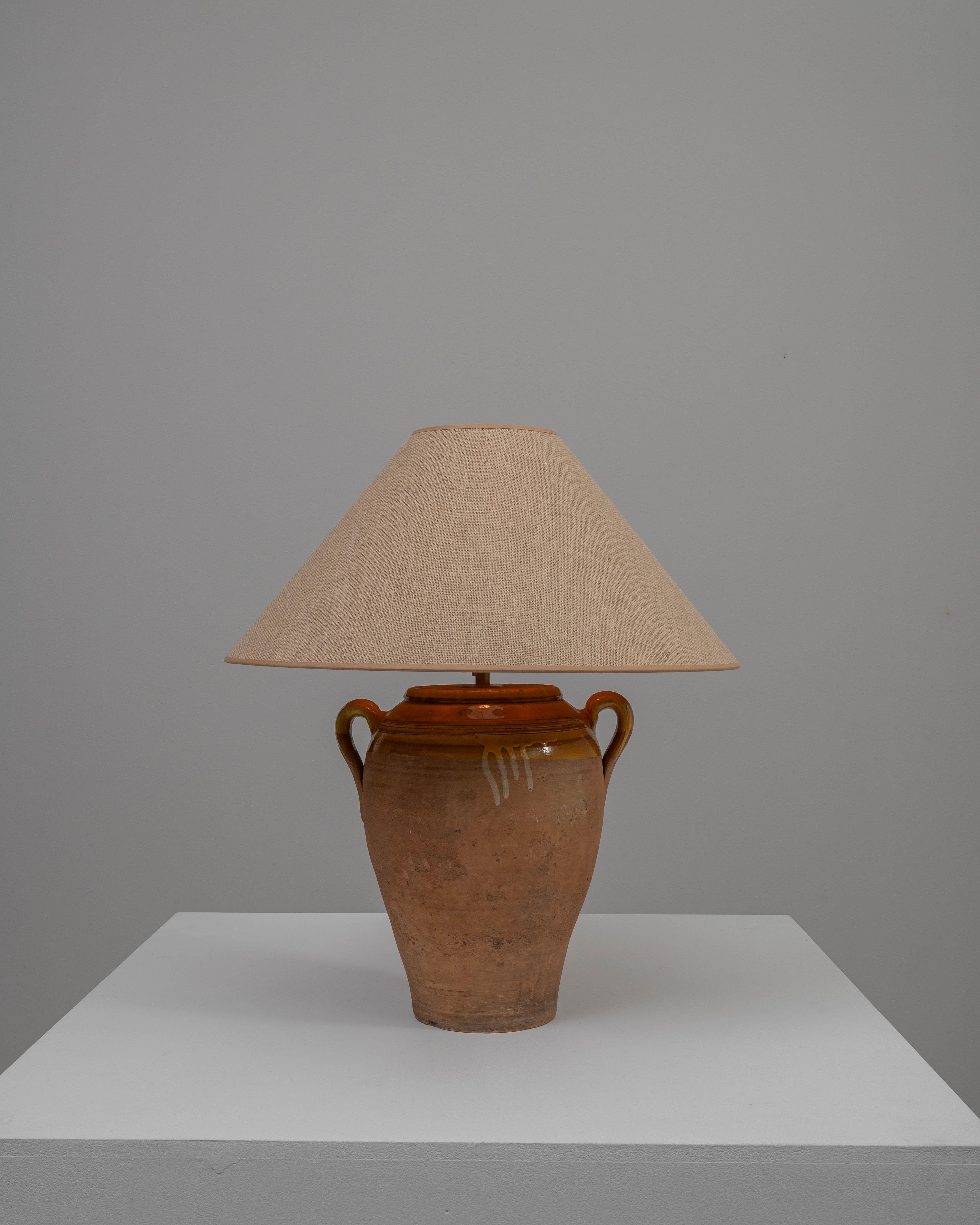 This 20th Century German Ceramic Table Lamp exudes a certain provincial charm with its antique pottery style. The terracotta-hued ceramic base, reminiscent of an amphora used in ancient times, features a gracefully aged patina and rustic glaze drips