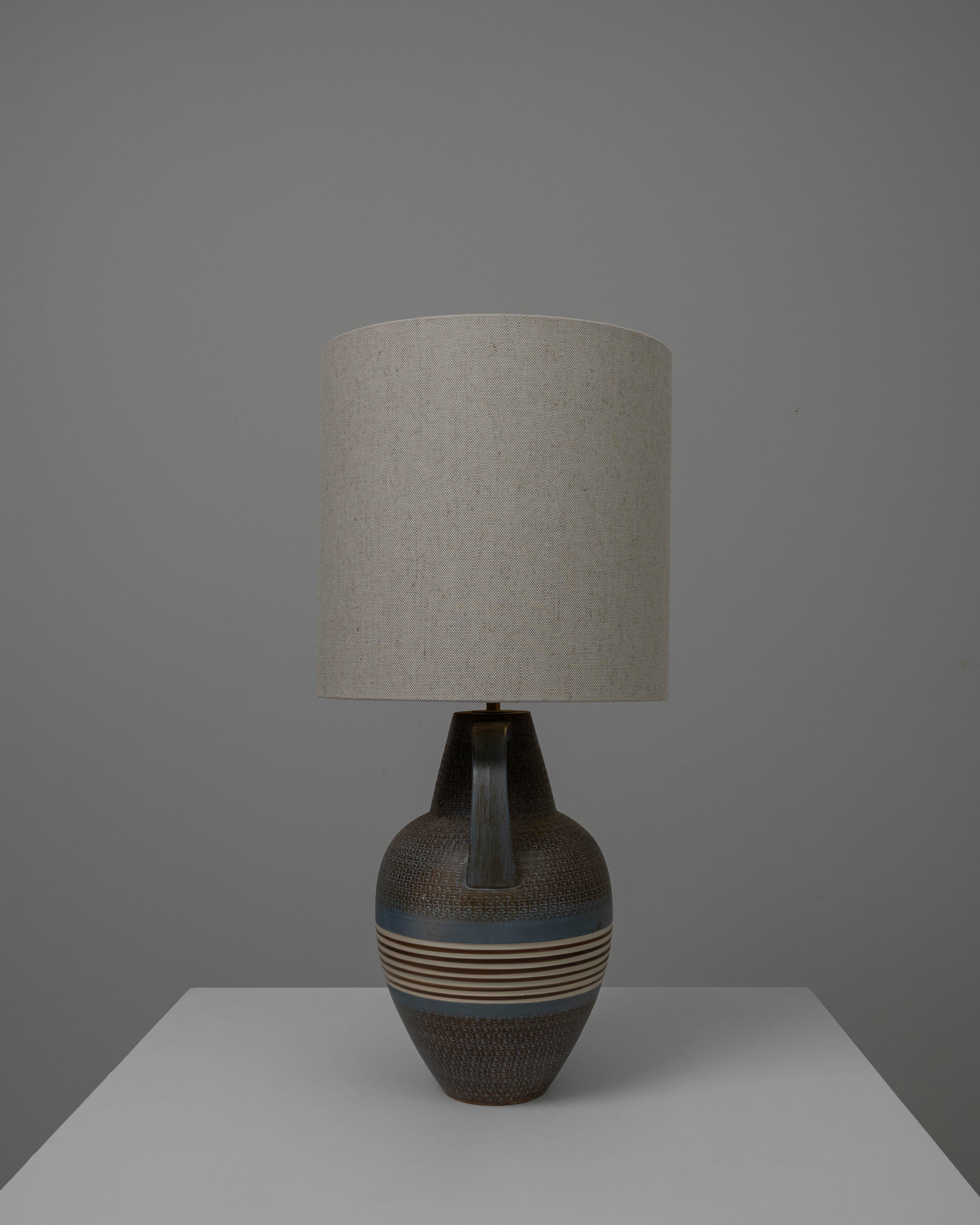 This 20th Century German Ceramic Table Lamp encapsulates the earthy, textural appeal of the era's artisanal design. The base, with its robust form and a handle for a touch of old-world charm, features a deep, variegated glaze of browns and greys,