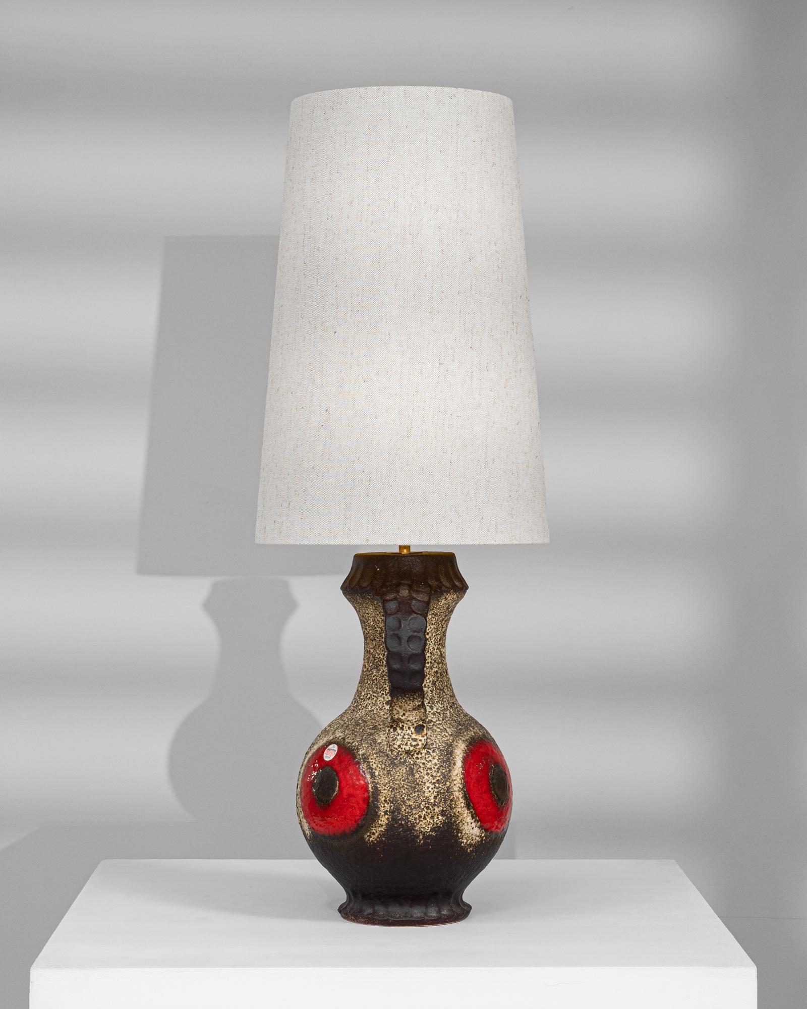 Cette lampe de table en céramique allemande du XXe siècle allie un design audacieux à un savoir-faire artisanal. Sa base en forme de cruche présente une finition sable texturée, accentuée par des motifs circulaires rouges frappants soulignés de