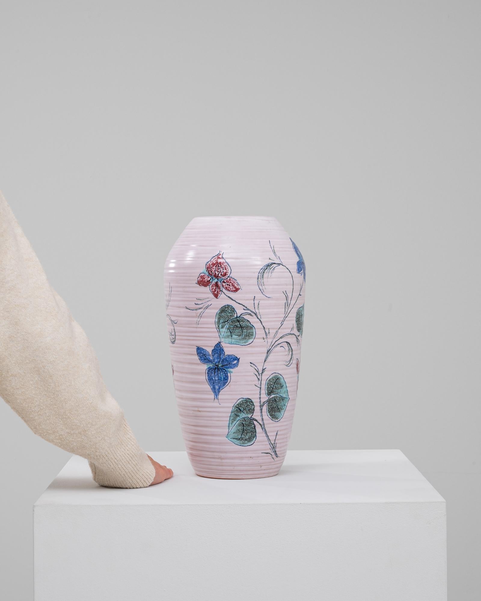 Este delicado jarrón de cerámica alemana del siglo XX es un encantador ejemplo de arte pintado a mano. Su suave cuerpo rosa está adornado con intrincados motivos florales en vibrantes tonos azules, verdes y rojos, que añaden un toque de elegancia y