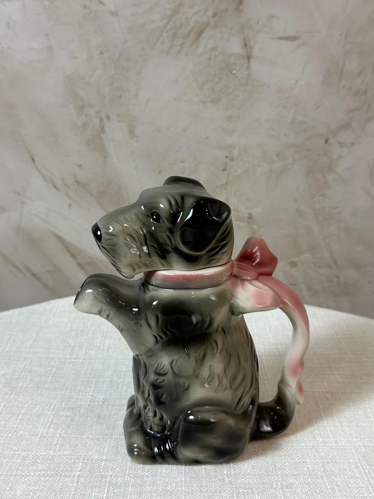 Llamativa y decorativa tetera de porcelana que representa a un perro sentado con un pa�ñuelo rosa alrededor del cuello.
Esta encantadora pieza data de los años 50 y procede probablemente de una manufactura alemana, famosa por sus creaciones